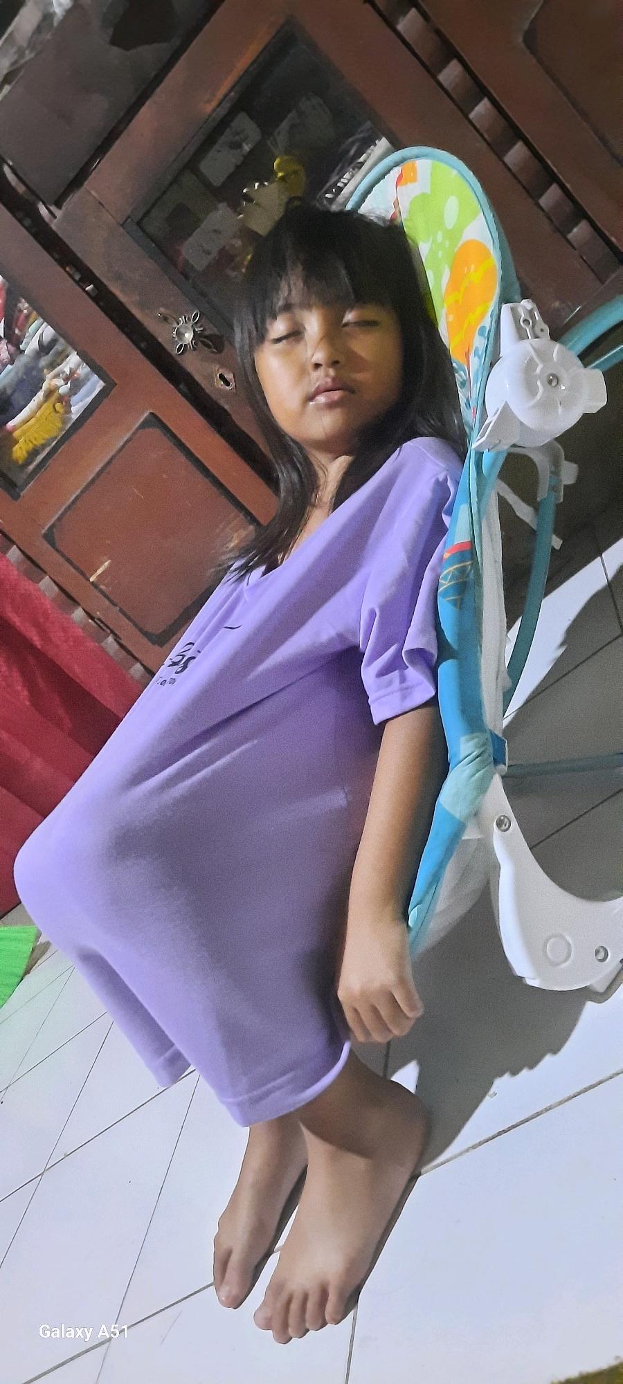 A young girl with closed eyes and dark hair is sitting on the floor in what appears to be a residential indoor environment in Jatitengah, Indonesia. She is wearing an oversized light purple t-shirt. Next to her is a baby swing with a colorful patterned seat and white plastic components. In the background, a wooden cabinet with glass doors is visible, containing various items. The floor is tiled with white squares. The lighting suggests it is nighttime.