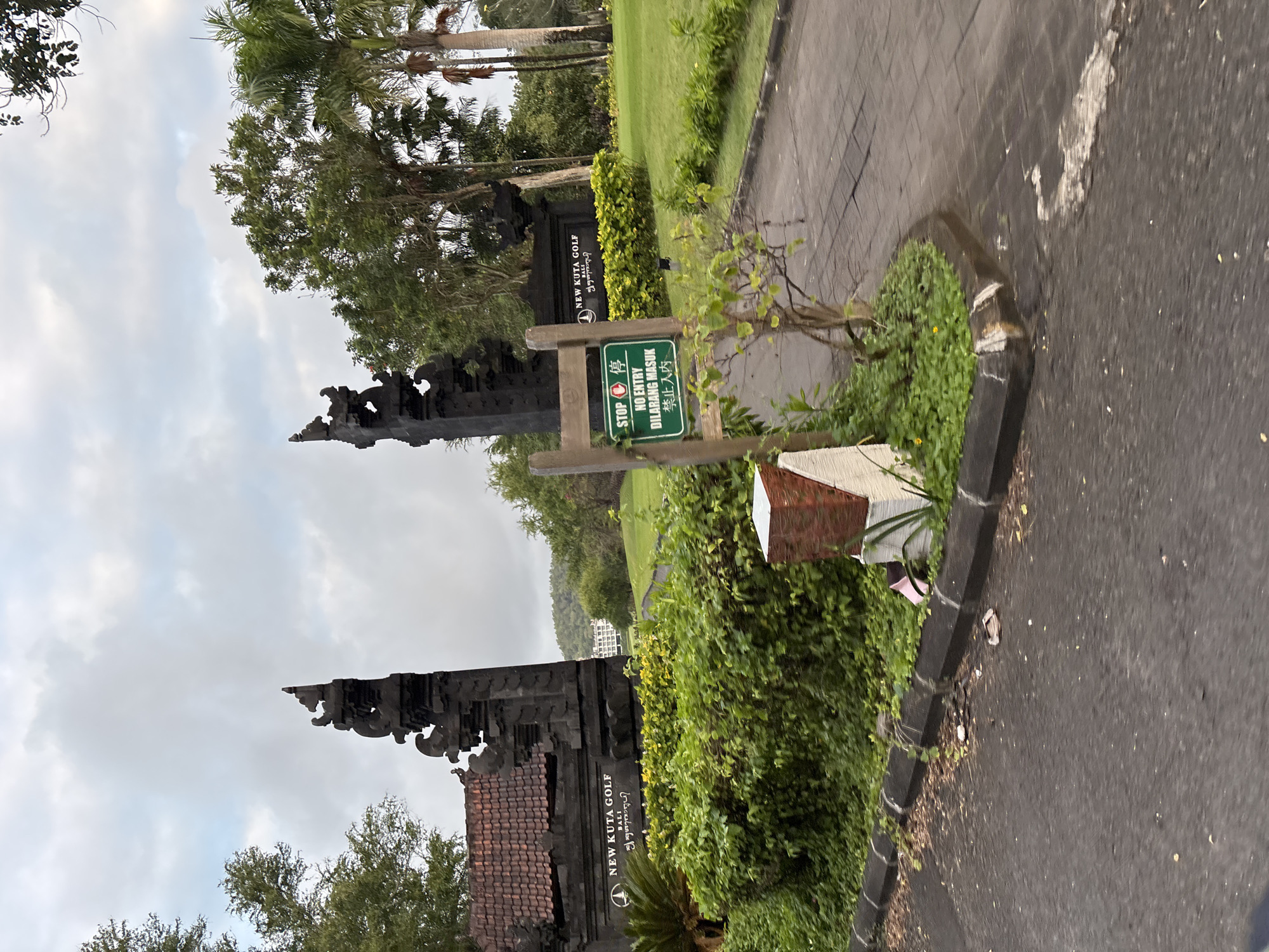 The image captures an outdoor scene at the entrance of New Kuta Golf in Kuta Selatan, Indonesia. The entrance features traditional Balinese gate architecture, with ornate stone carvings and a tiled roof on the left structure. To the right of the entrance, a paved pathway leads into the golf course, flanked by lush green grass and trees. A prominent sign is visible, displaying "STOP" in English and Chinese characters, followed by "NO ENTRY" in English and Malay. Further down, a building complex with white walls and a dark roof is visible in the background, nestled amidst rolling green hills. The sky above is overcast with diffused sunlight. Several small plants and bushes are strategically placed around the entrance, enhancing the natural beauty of the location.