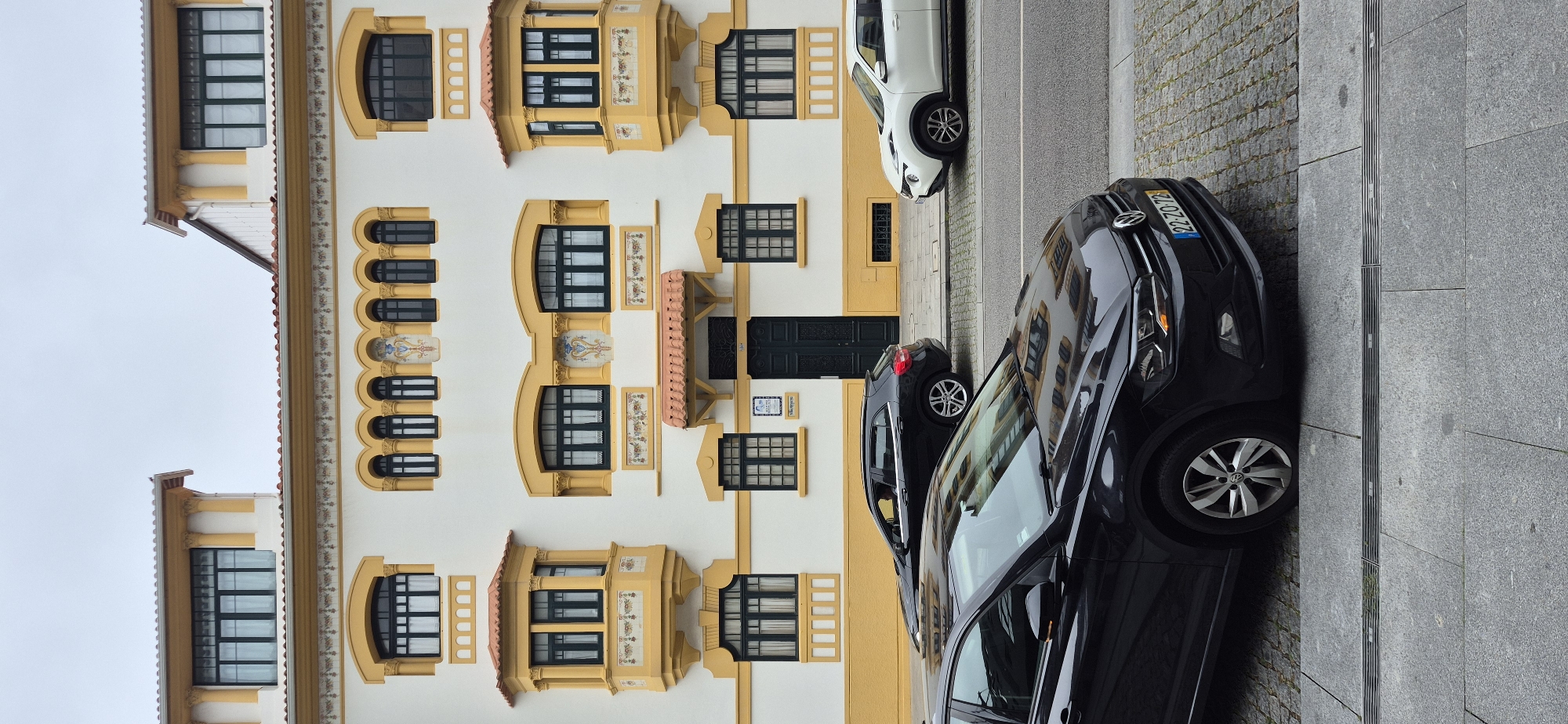 This image captures a street scene in Póvoa de Varzim, Portugal, featuring a white building with yellow architectural accents and several parked cars. The building, characterized by its ornate windows, bay windows, and decorative frieze, is a prominent element. In the foreground, a black Volkswagen Polo is parked at an angle on a sidewalk paved with large grey tiles and cobblestones. Behind it, a black SUV is parked further down the street, and a white Nissan Juke is parked on the right side of the image. The street is paved with asphalt. The sky is overcast.