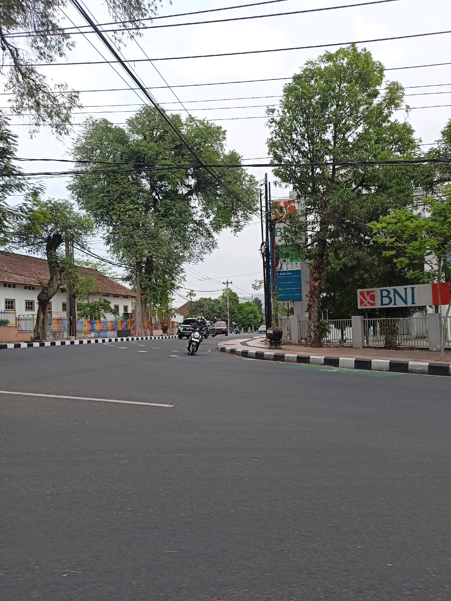 The image captures a street scene in Magelang, Indonesia, featuring a paved road with a white lane marking. On the left, a white building with a red-tiled roof and a colorful striped fence is visible, bordered by mature trees with lush green foliage. A motorcycle with a rider wearing a dark helmet is in motion on the road, heading towards the right side of the frame. Further down the road on the right, a building with a prominent BNI (Bank Negara Indonesia) logo is situated, with a metal fence in front of it. Several other trees with dense green leaves line the street, and overhead, a network of utility wires is strung across the sky, which is overcast. In the background, other vehicles and buildings can be observed, suggesting a typical urban environment.