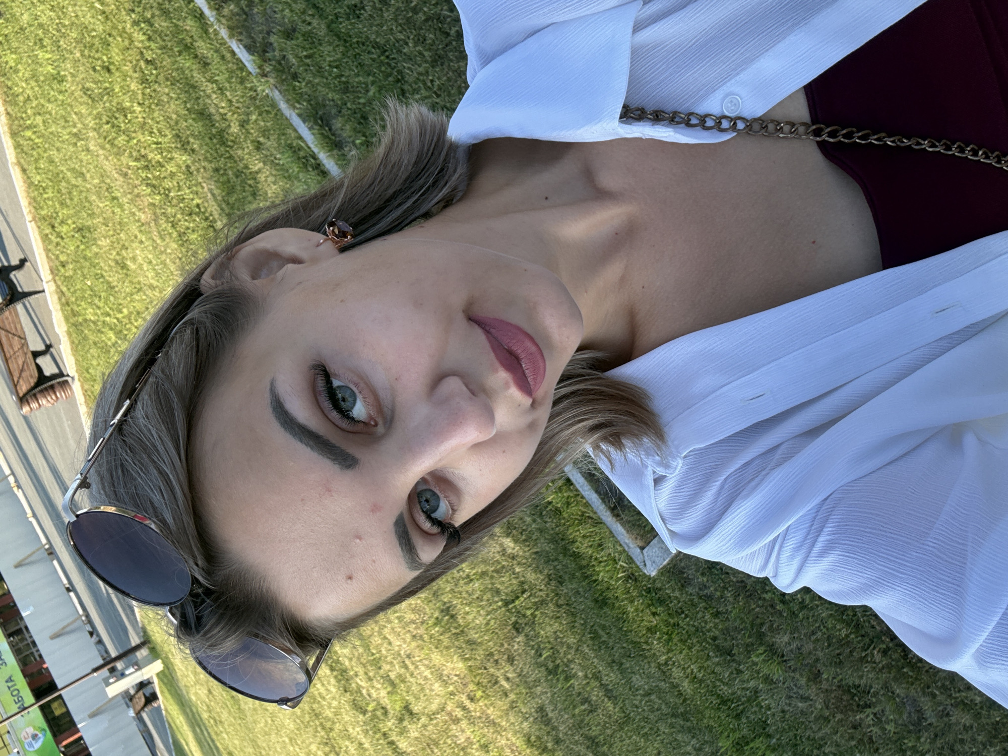 A woman with blonde hair and blue eyes is taking a selfie outdoors. She is wearing a white shirt and sunglasses are perched on her head. Her makeup includes mascara and lipstick. She is also wearing a small earring in her right ear. The background shows a grassy area with a pathway and some benches, suggesting a park or public space. In the distance, there appears to be a building with signage, possibly in Yuzhno-Sakhalinsk, Russia.