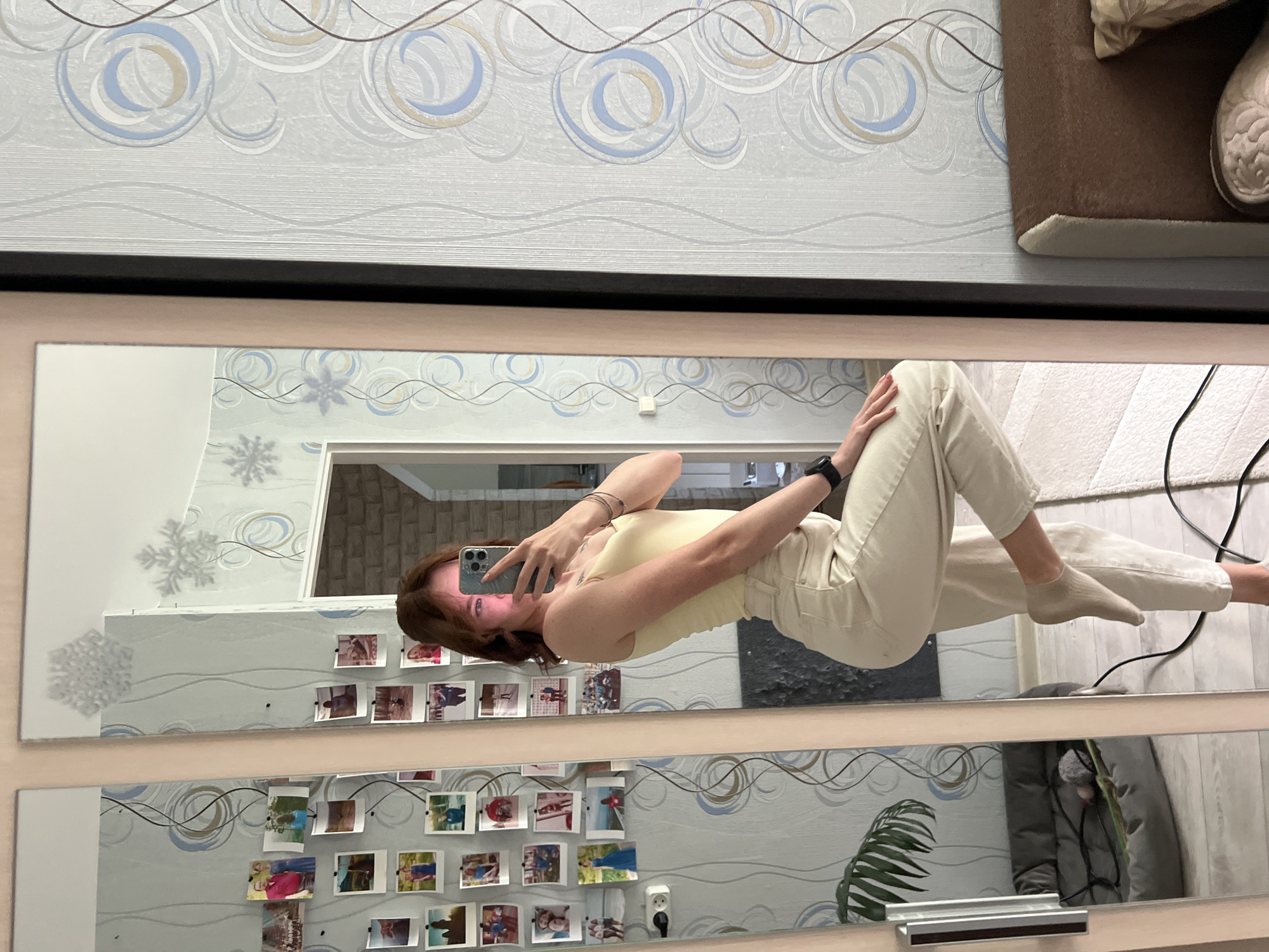 An indoor scene in Chaikovsky, Russia, features a young woman taking a mirror selfie. The image, presented rotated 90 degrees clockwise, depicts the woman reflected in a large, rectangular mirror with a light brown frame and black inner border.
The woman has short, reddish-brown hair and is wearing a light yellow sleeveless top and light beige or cream-colored trousers. She holds a light blue/green smartphone with multiple camera lenses, looking at the screen as she captures her reflection. A black watch is visible on her left wrist. She is standing with her legs slightly bent, wearing light-colored socks, with a light-colored carpet or rug visible on the floor in front of her.
The wall visible in the mirror's reflection, behind the woman, is adorned with light grey wallpaper featuring an abstract pattern of blue, beige, and dark brown swirling lines, along with stylized snowflake motifs. Numerous small, square instant photos are taped to this wall, showcasing various people and scenes, adding a personal touch to the decor.
In the reflected view, a white power outlet with two sockets is seen on the wall, along with a green leafy plant. Outside the mirror's frame, in the top portion of the original image, more of the patterned wallpaper is visible, along with a glimpse of dark brown furniture, possibly a sofa or a headboard, and a beige pillow or cushion. No discernible text is visible.