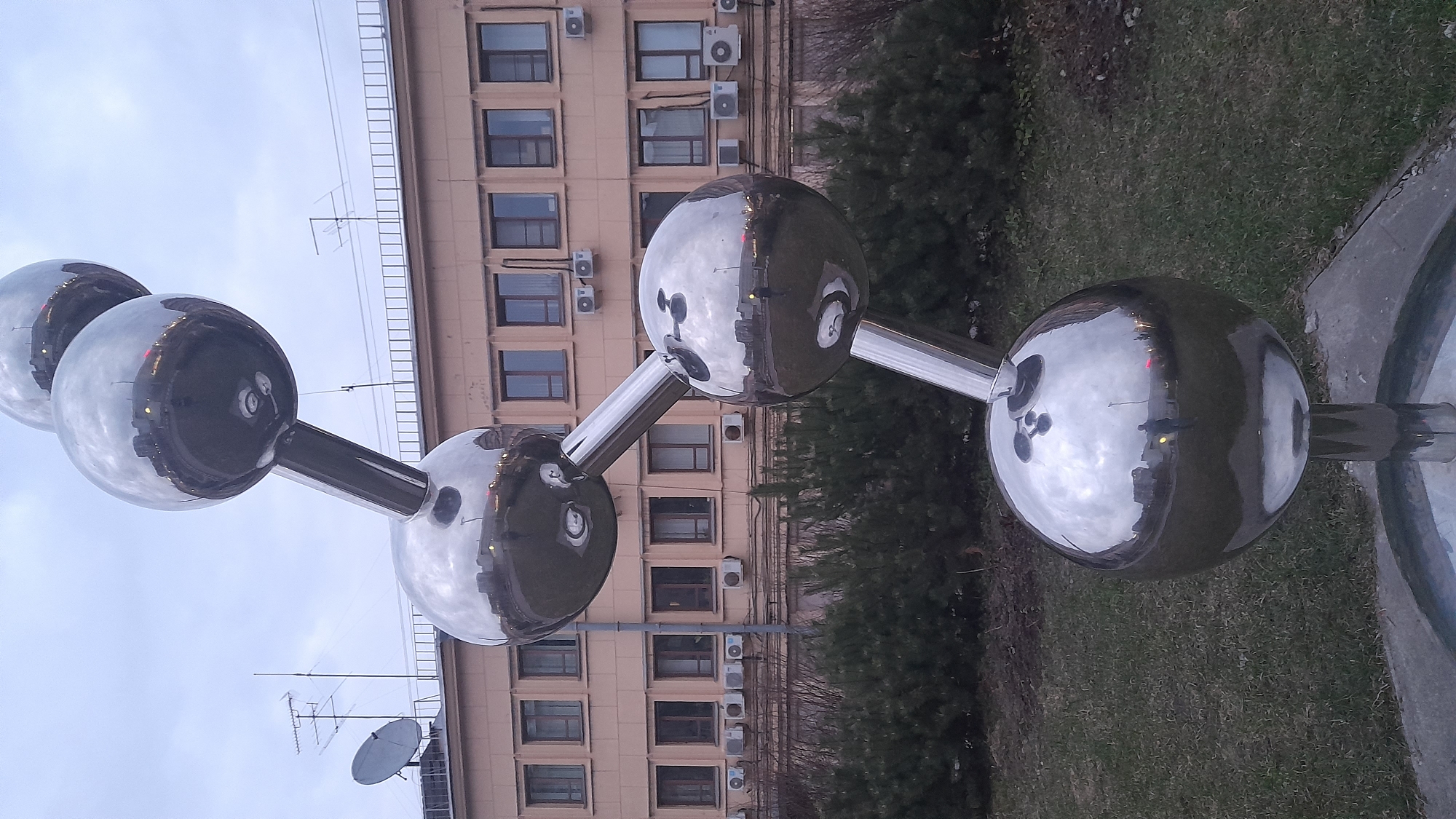 A low-angle, sideways view shows a shiny metal sculpture of connected spheres resembling a molecule, positioned outdoors in front of a light-tan building in Saint Petersburg, Russia. The sculpture features four reflective spheres of varying sizes connected by metal bars. Reflections in the spheres show sky and urban elements. The building behind the sculpture has multiple floors with a repeated pattern of windows and air conditioning units. Patches of greenery surround the base of the sculpture. The sky is overcast. An array of antennas and a satellite dish are above the sculpture.