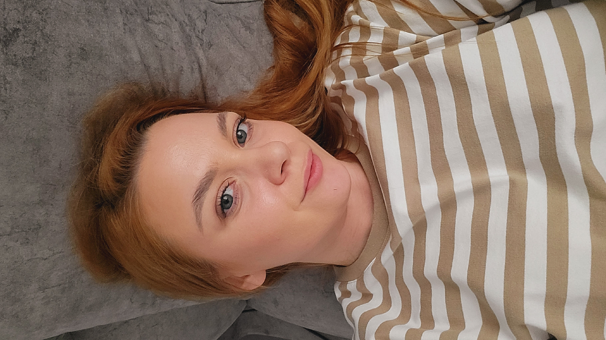 A medium close-up, high-angle shot features a young woman lying down, looking directly towards the camera with a gentle, friendly expression. Her head is tilted to the right, resting on a soft, textured grey surface, likely a sofa or bed. She has fair skin, light-colored eyes (appearing blue-grey or green), and reddish-brown hair that frames her face and spreads around her head. Her eyelashes are prominent, and she has well-defined eyebrows. She wears a long-sleeved shirt with alternating vertical stripes in beige or light brown and white. The overall impression is one of calm and relaxation, within an indoor setting. There is no visible text. The location context for this image is Chaikovsky, Russia.