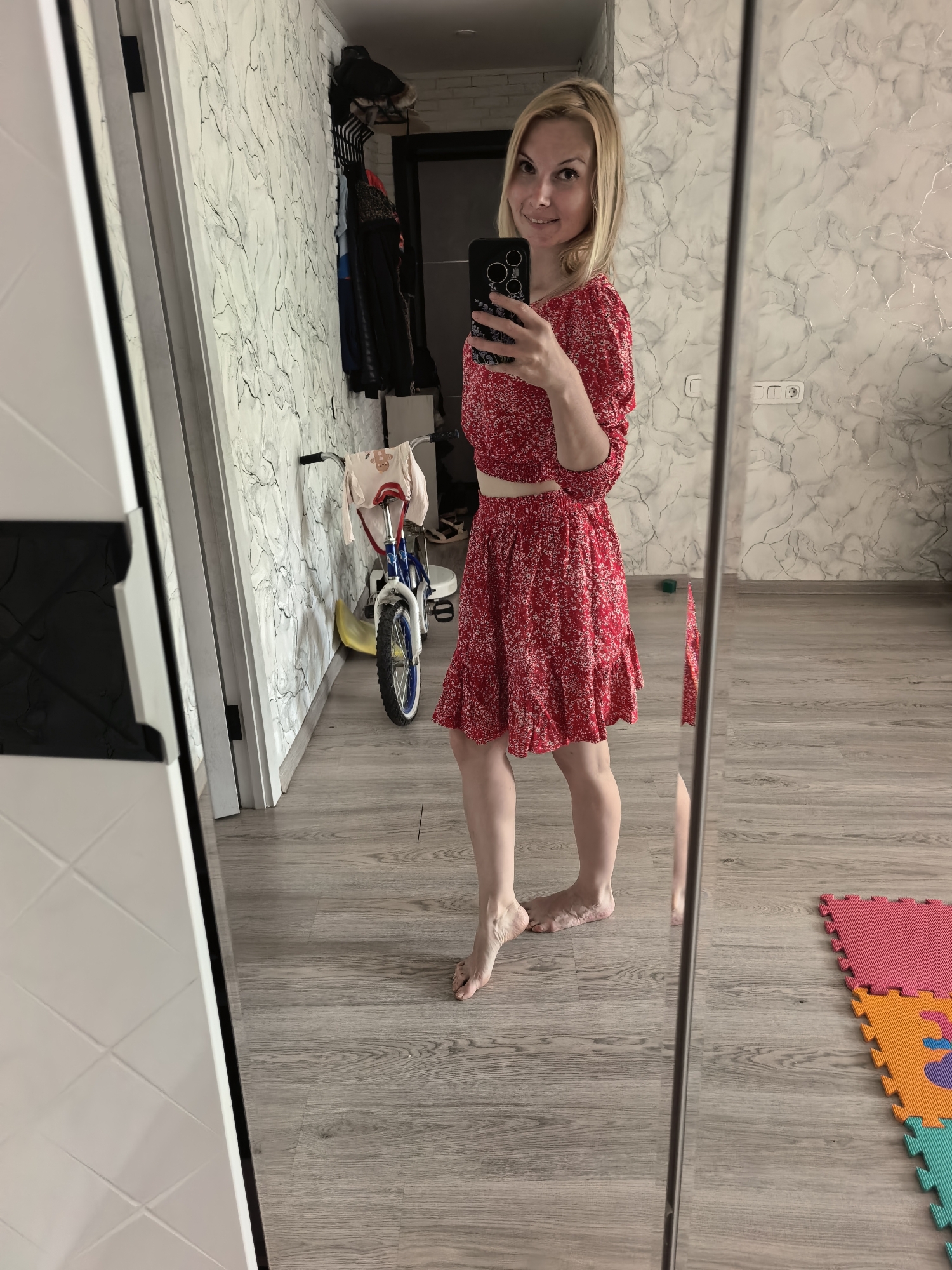 A woman is taking a mirror selfie indoors. She is fair-skinned with blonde hair, wearing a red floral print two-piece outfit. She is holding a smartphone with multiple rear lenses.
The interior has light-colored hardwood floors. In the background is a child's bicycle leaning against the wall, a clothing rack with coats and jackets. On the floor are interlocking foam play mats in various colors.
The location appears to be a residential space in Sterlitamak, Russia, based on architectural styles and the presence of domestic items.