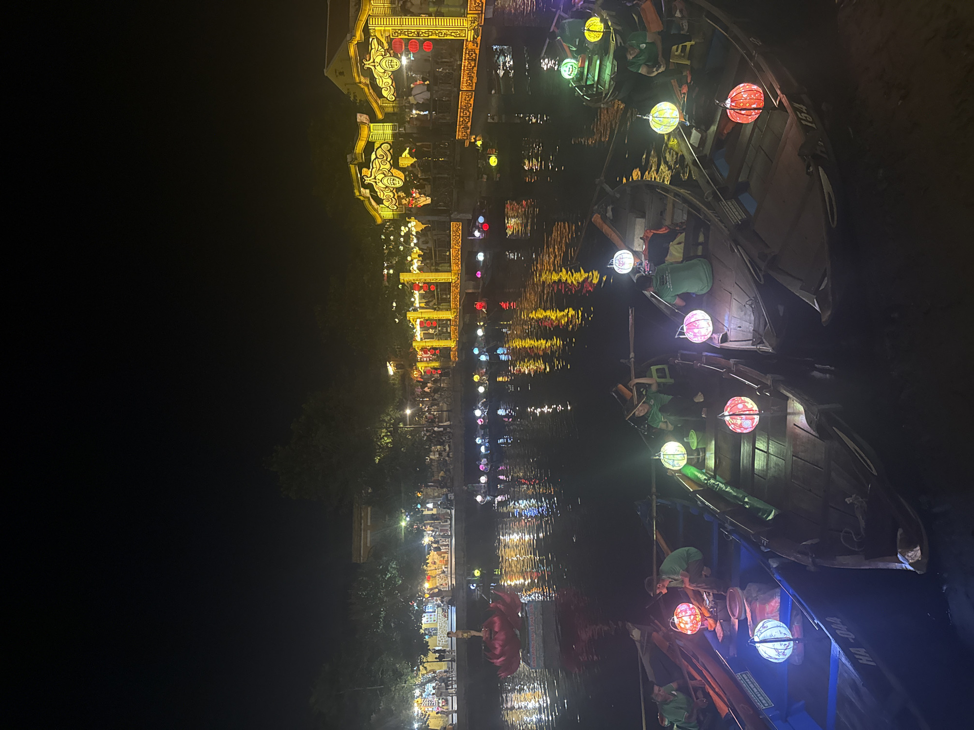 Captured at night, the image portrays a river scene in Unknown, Unknown. Boats, adorned with illuminated lanterns, are a primary focus. The light from the lanterns is also reflected in the water. Buildings are visible on the riverbank, brightly lit with decorative lights, adding to the festive atmosphere. Pedestrians line the riverfront.