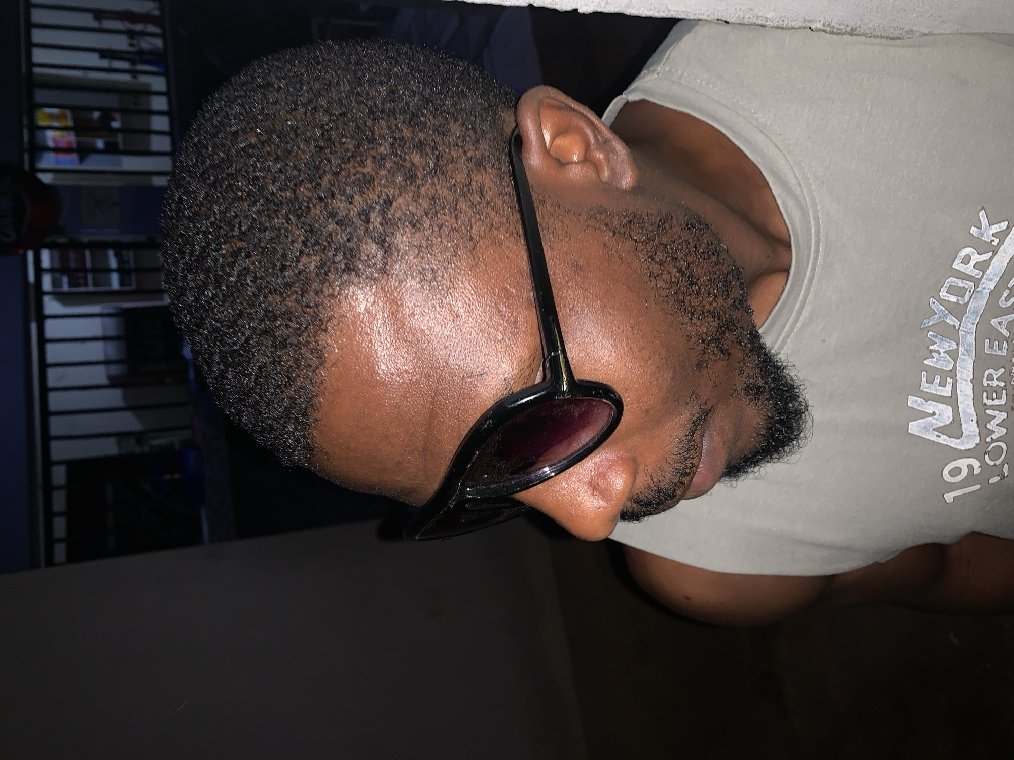 A medium-close shot shows a dark-skinned man with a short, cropped haircut and a beard. He is wearing sunglasses with dark lenses and a light-colored t-shirt with the word "NEWYORK" printed on it. The photo is taken indoors and the lighting is uneven, with some areas appearing darker than others. A rack or shelving unit is faintly visible in the background. No geolocation information could be located.