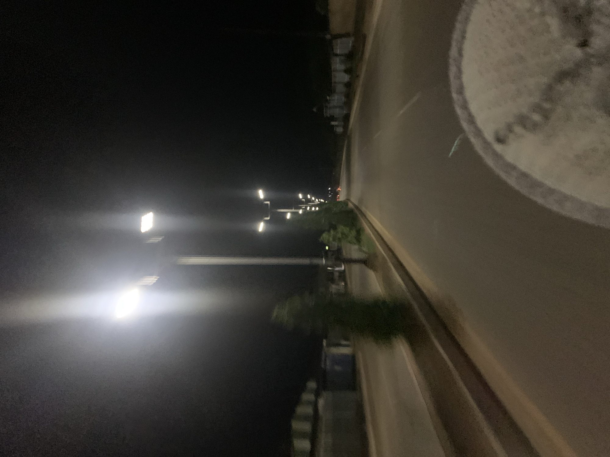 This image depicts a nighttime scene on a multi-lane asphalt road in the city of New Stadium, Nigeria. The photograph appears to be taken from a moving vehicle, evidenced by motion blur in certain areas. The sky is dark, indicating a lack of natural light.
The road itself is illuminated by numerous tall, metallic streetlights, predominantly on the left side, each equipped with multiple bright, white LED fixtures. The closest streetlight on the left exhibits significant lens flare and vertical light streaking. A raised median or curb separates the road from a strip of vegetation, where several blurry green trees or bushes are visible, including one prominent conical shape in the mid-ground on the left. Beyond the vegetation on the left, indistinct, low-rise structures or buildings with flat roofs can be observed, some appearing light grey or white, with one section showing a blue hue.
On the right side of the road, further down, additional streetlights are visible, along with a darker landscape comprising vegetation and what appears to be a low wall or embankment, and a white fence-like structure in the distance. The road surface reflects the artificial light, showing variations in grey and brown tones.
In the bottom right foreground, a large, blurry, circular object with a textured, light-grey surface and a darker rim is partially visible, consistent with a vehicle's interior component like a tire or steering wheel. A faint, glowing blue-green streak is present on the road surface directly above this object. No people or overt actions are discernible in the frame. The environment is an urban or semi-urban roadway at night.