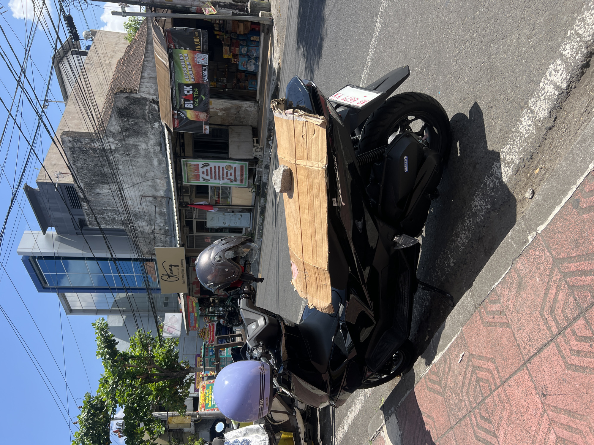 A black scooter is parked on the side of a street in Yogyakarta, Indonesia. The scooter has two helmets on it, one lavender and one dark gray. A piece of cardboard is secured to the back of the scooter with a rock on top. In the background, there are several buildings, including a modern building with blue glass windows and a more traditional building with a tiled roof. There are also several power lines running overhead. A sidewalk with red patterned tiles is visible in the foreground.