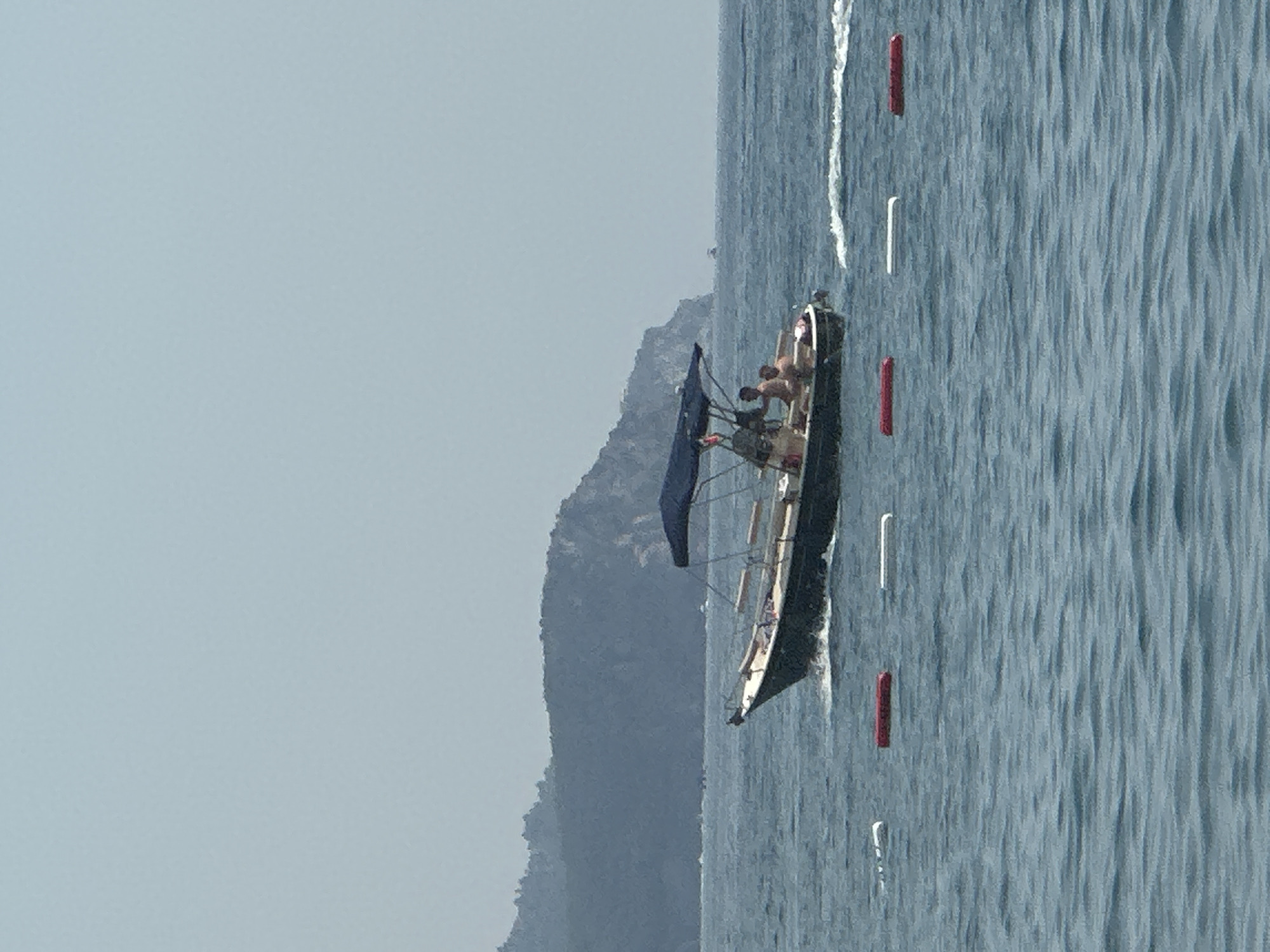 A dark-hulled motorboat with a blue canopy is traveling through calm, blue water. Three individuals, two appearing to be adult males, are seated on the boat. The boat is leaving a white wake. In the background, a hazy, tree-covered mountainous coastline is visible under a pale blue sky. Several buoys, some red and some white, are floating in the water in the foreground. The scene appears to be set on the coast of Boreti, Montenegro, based on the geographical features.
