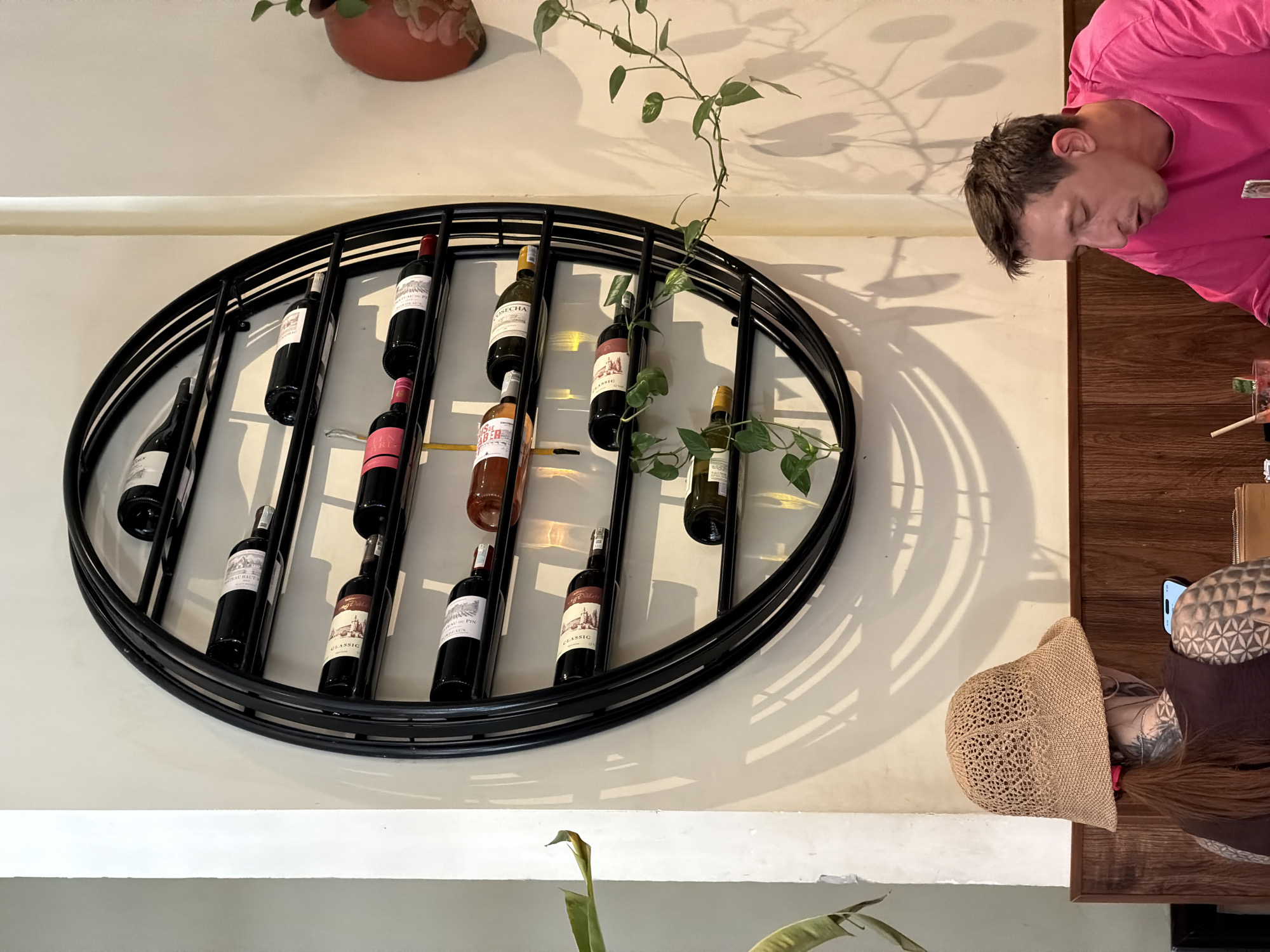 In Nha Trang Ward, Vietnam, a circular black metal wine rack is mounted on a cream-colored wall. The rack displays multiple bottles of wine on horizontal shelves, some of which are partially obscured by shadows cast by the bottles and a nearby plant. In the foreground, a person wearing a woven hat and a dark top with visible tattoos is seated, looking at a phone. To their right, a man in a bright pink shirt is also seated, looking down. The lower portion of the image reveals a wooden wall paneling.