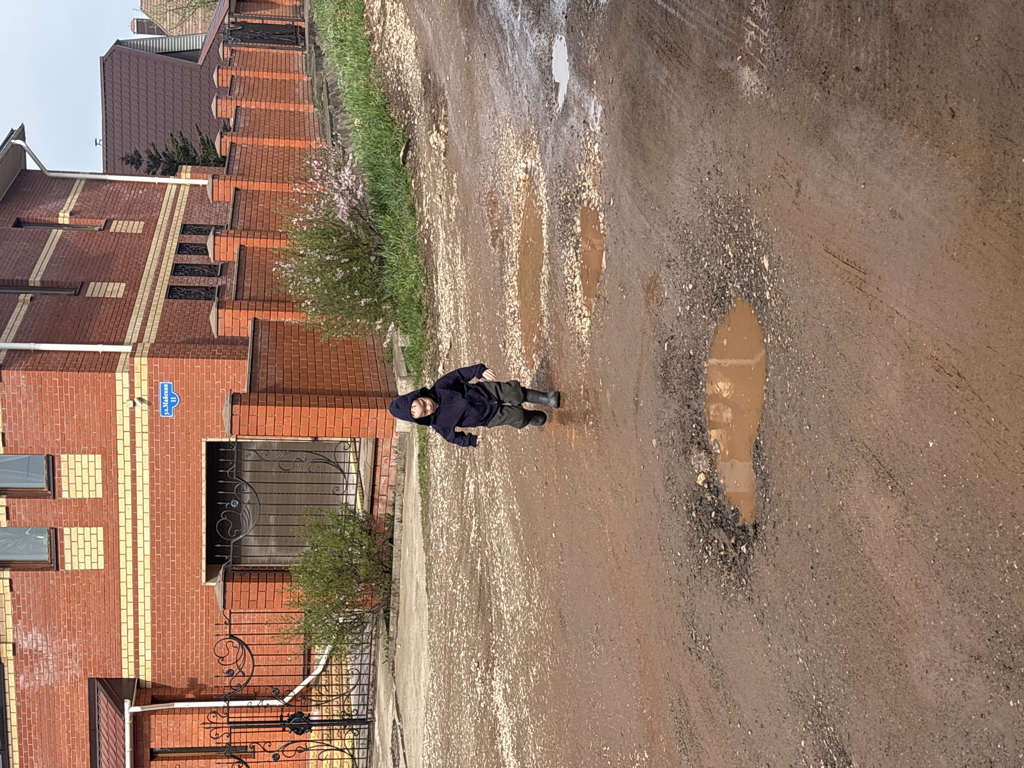 A young person wearing a dark-colored hooded jacket and dark trousers, with black boots, is lying on a muddy, potholed dirt road in Ферма, Russia. The road is adjacent to a red brick building with a black wrought-iron gate and window grating. A street sign reading "ул. Маяковски 11" (Mayakovsky Street 11) is affixed to the building. A narrow strip of grass and blooming vegetation separates the road from the building. Puddles of muddy water are visible in the road. A partial view of another building with a brown tiled roof is visible in the upper left corner. The sky is overcast.