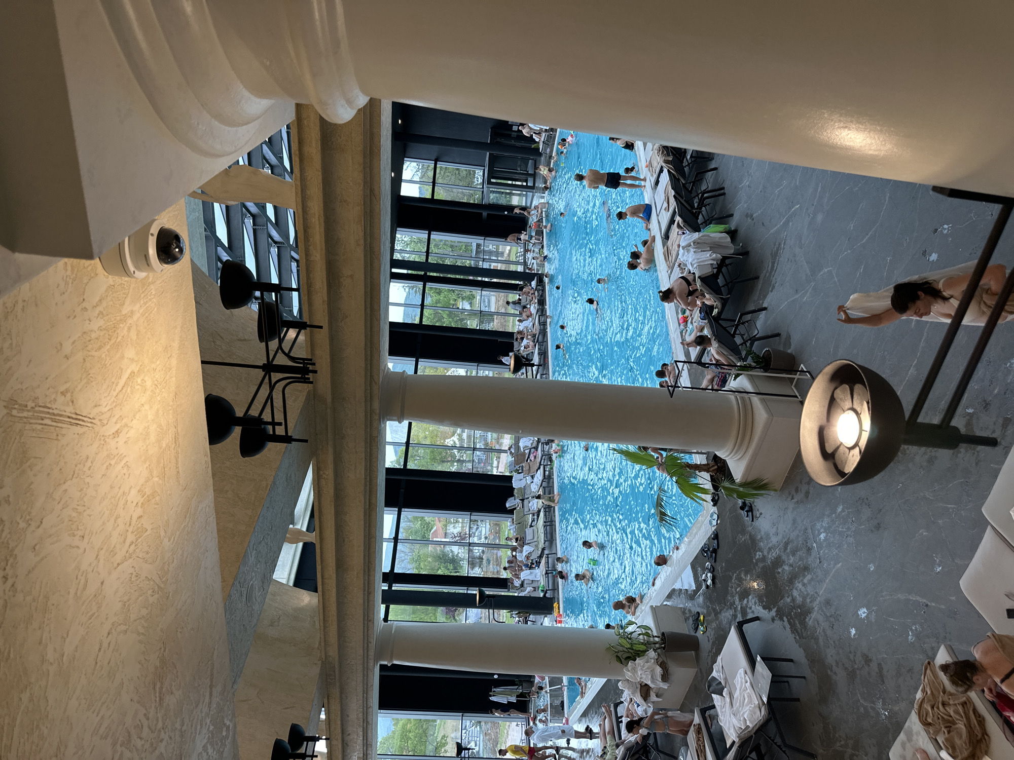 Here's a detailed description of the image:
**Scene Overview:**
The image captures an indoor swimming pool scene, possibly within a spa or recreational facility. The perspective appears to be from an elevated position, overlooking the pool and its surroundings.
**Location Details:**
The city is Kazanlak, Bulgaria.
**People:**
Numerous people are present in the scene. Some are swimming in the pool, while others are lounging on chairs alongside the pool. The individuals appear to be of various ages and are dressed in swimwear or wrapped in towels. There are women sunbathing.
**Subjects and Objects:**
* **Pool:** A large indoor swimming pool filled with clear blue water.
* **Chairs:** Numerous lounge chairs are arranged along the pool's edge, some occupied by people.
* **Columns:** Large, decorative columns are visible in the foreground, adding to the architectural detail of the space.
* **Windows:** Large windows are present in the background, allowing natural light to enter the area. Trees and foliage are visible outside the windows.
* **Lights:** The scene is illuminated by both natural light and artificial light sources, including overhead lighting fixtures.
* **Security Camera:** A security camera is mounted on the ceiling, indicating a surveillance system is in place.
* **Lamps:** Some lamps are included in the foreground area.
* **Plants:** Some plants are located in the area.
**Actions and Interactions:**
* People are swimming, lounging, and interacting with each other.
* Some individuals are drying off with towels or relaxing in the lounge chairs.
**Environment:**
* **Architecture:** The location features a sophisticated architectural design, with columns, large windows, and decorative elements.
* **Lighting:** The combination of natural and artificial lighting creates a bright and inviting atmosphere.
* **Overall Impression:** The scene suggests a relaxed and recreational environment, likely within a spa or leisure facility.