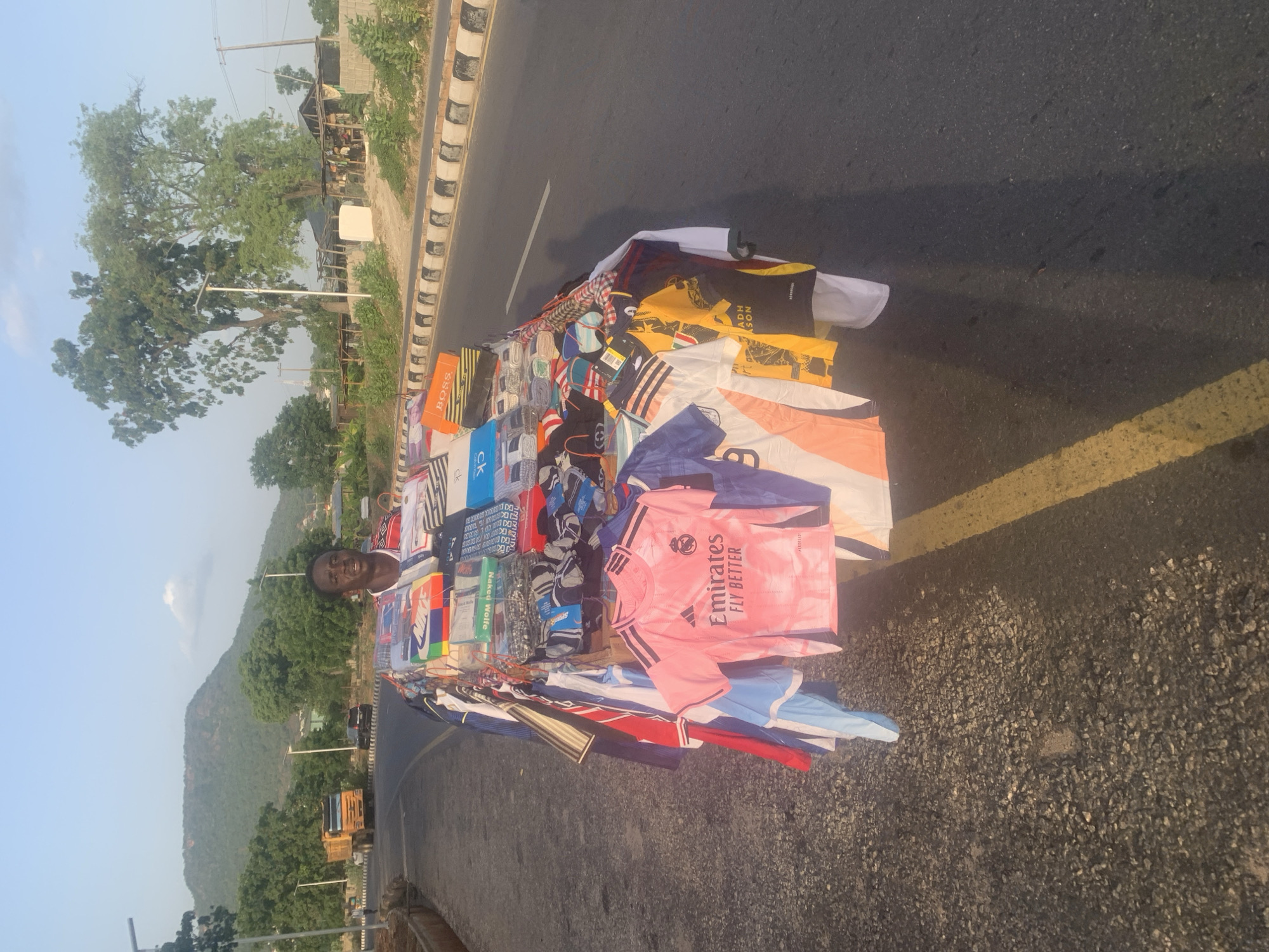 The image shows a street vendor in Zango-ndaji, Nigeria, displaying clothing and accessories for sale on a table set up on the side of a road. The vendor, a dark-skinned man, is visible behind the merchandise. The items for sale include shirts, socks, underwear and boxes from name brand companies. The road is asphalt, and a yellow line marks a lane divider. In the background are trees, buildings, and a mountain range. The sky is light blue, suggesting it is daytime.