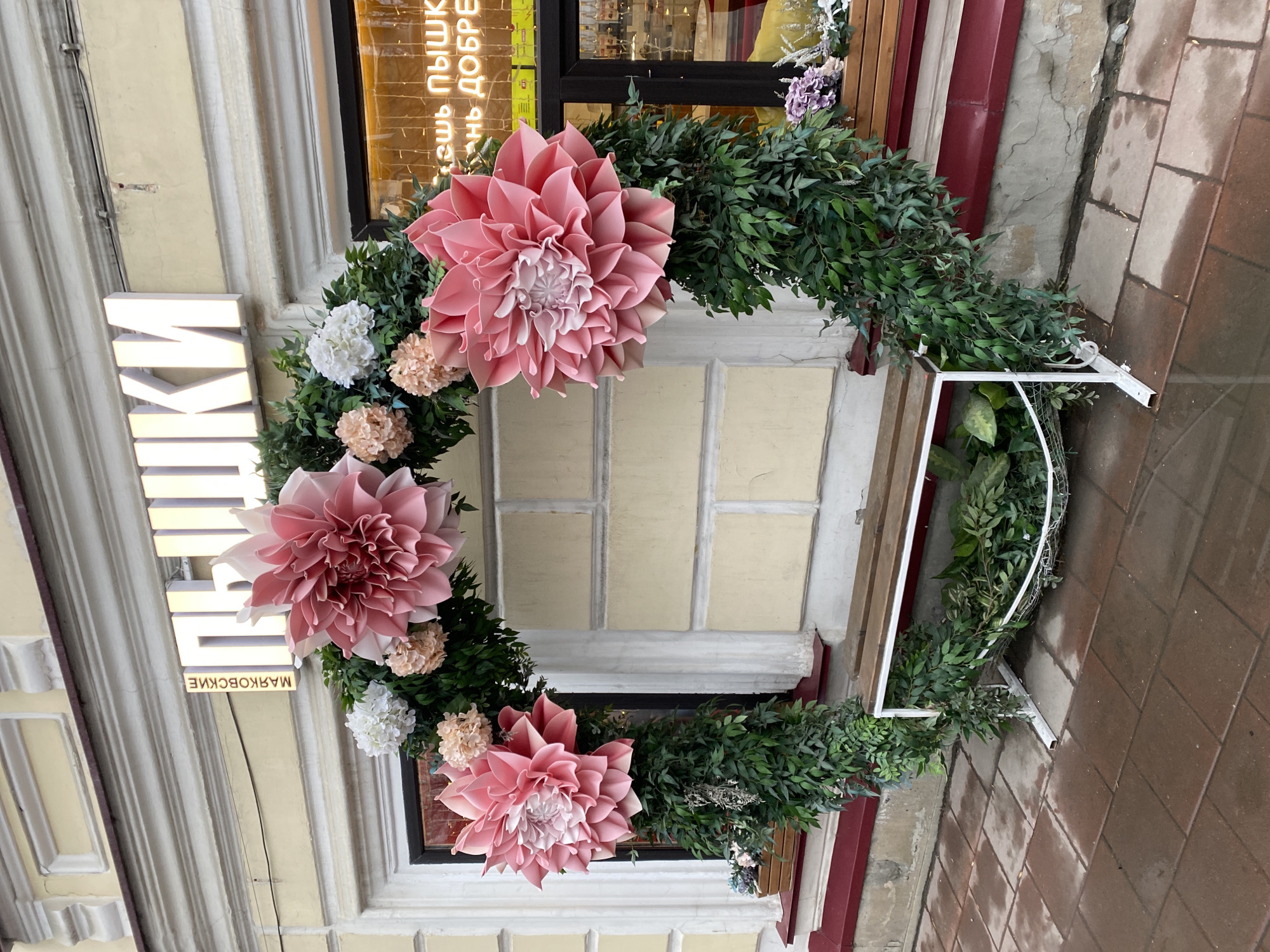 A large, semi-circular floral and greenery arch decorates the facade of an old building, likely a storefront, in Yekaterinburg, Russia. The arch is composed primarily of dense, dark green foliage, accented with several oversized, light pink paper or fabric flowers resembling dahlias, interspersed with smaller clusters of white and pale peach-colored artificial blooms. The decorative arch stands on a white metal frame, with some greenery cascading into a trough-like base at the bottom right.
The building facade is light-colored, possibly cream or pale yellow, with intricate architectural details including pilasters and molding around the windows. The storefront features a dark-framed window or door, through which interior lighting is visible, casting a warm glow. Reflections in the window reveal Cyrillic text, reading "...ешь ПЫШКА...нь ДОБРЕ", suggesting a slogan related to food, possibly "Ешь пышки, добрый день!" (Eat doughnuts, good day!).
To the left of the main floral display, a vertical, illuminated white sign with raised Cyrillic letters is mounted on the building. The visible text on this sign reads "ЛАВКИ МАЯКОВСКИЕ", which translates to "Mayakovsky Shops" or "Mayakovsky's Stalls", likely indicating the name of the establishment or its location on a street named after Mayakovsky.
The sidewalk in front of the building is paved with reddish-brown tiles and appears wet in several areas, suggesting recent rain. A narrow, dark red vertical pipe or drain runs down the side of the building on the right. The overall impression is a vibrant and inviting decorative display against a historic urban backdrop, likely an entrance to a shop or cafe.