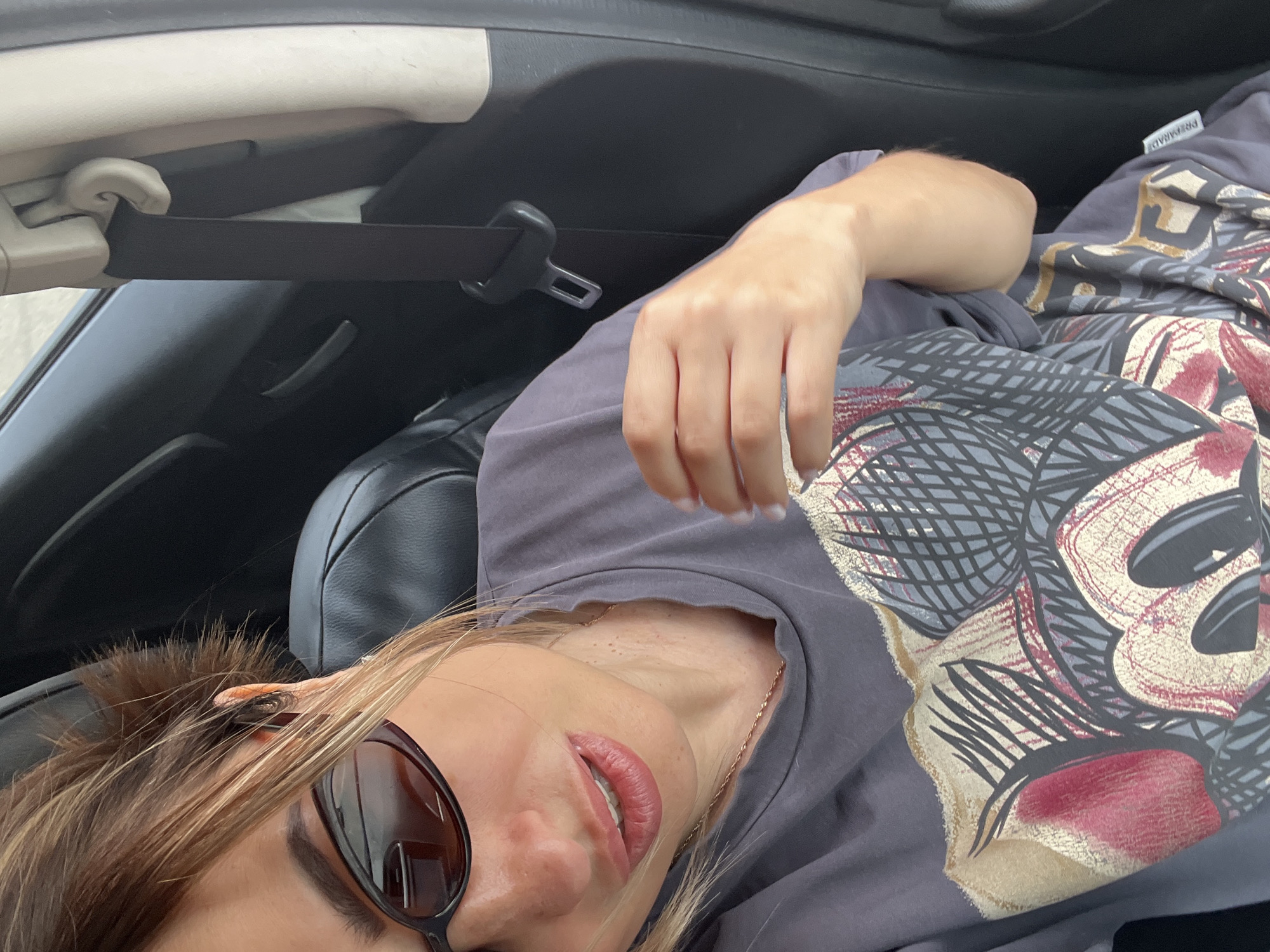 A woman with blonde hair pulled back in a messy bun, wearing dark-rimmed sunglasses and a grey t-shirt with a graphic print of Mickey Mouse, is shown from the chest up in the driver's seat of a car. Her mouth is slightly open, and her right hand is raised with fingers spread. The seatbelt is visible and buckled. The background is dark, suggesting the interior of a car, with a hint of a window in the upper left corner. The lighting suggests an outdoor, daytime setting.