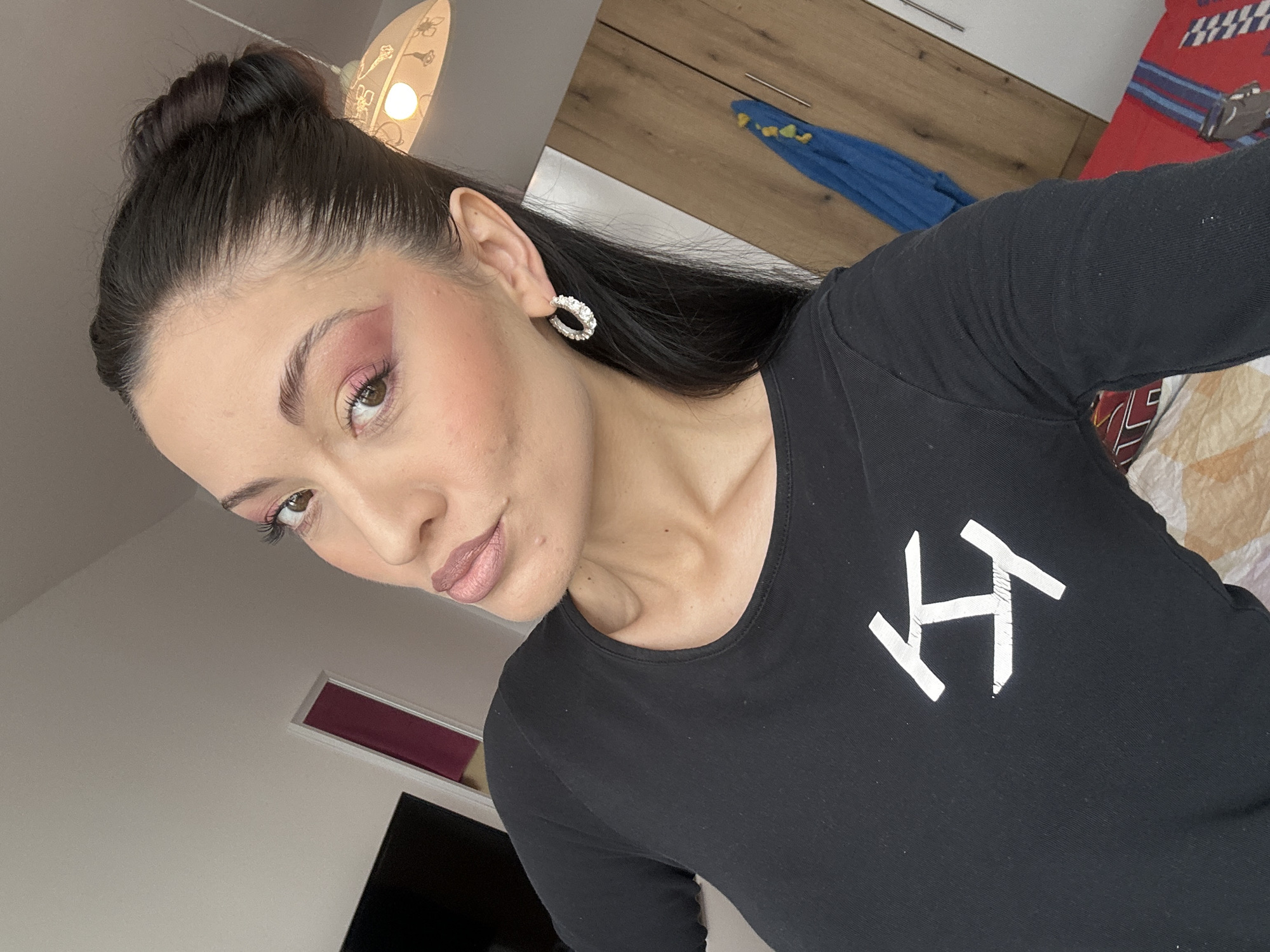 A young woman with brown hair in a high bun and pink eyeshadow is posing for a selfie indoors. She is wearing a black long-sleeved shirt with a white graphic on the chest and round earrings. The background shows a bedroom with a light fixture, wooden cabinets, a blue towel, and a red and blue patterned bedspread. There is also a glimpse of a window and a black TV screen. The image does not provide any information about the location being Burgas, Bulgaria.