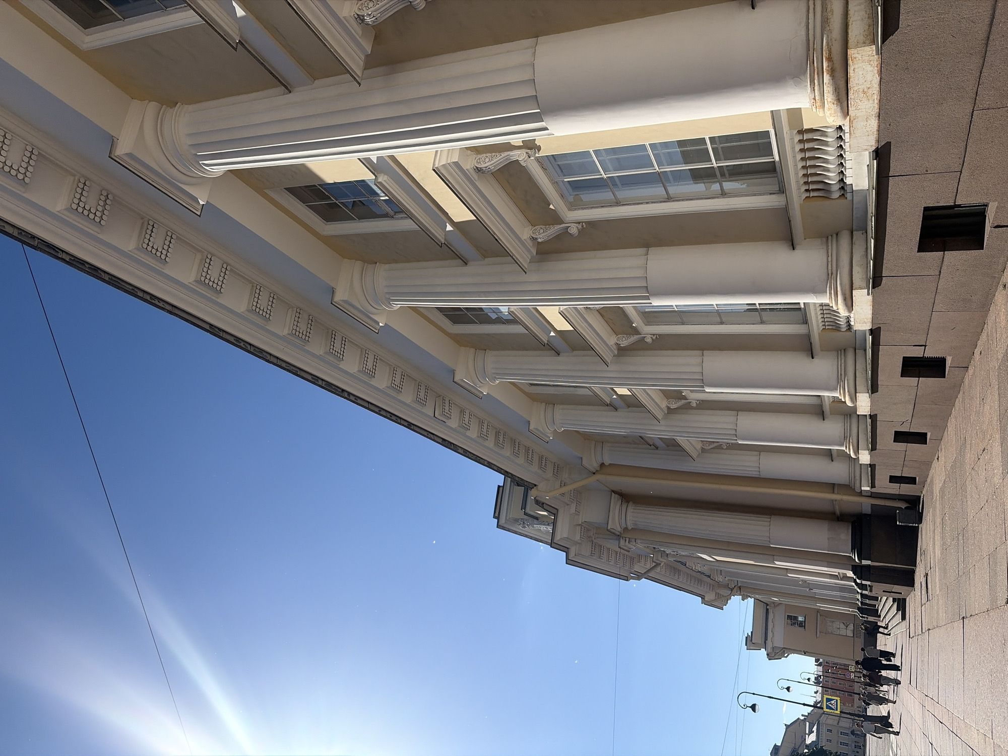 The image shows the facade of a grand classical building in Saint Petersburg, Russia, bathed in bright sunlight under a clear blue sky. The building features a long row of white Corinthian columns supporting a detailed cornice with decorative elements. Large, multi-paned windows are visible between the columns, reflecting the sky. The lower part of the building is clad in dark, textured stone. A paved sidewalk runs along the front of the building, and a street with lampposts and a pedestrian crossing sign is visible on the left. Several people are walking on the sidewalk, casting shadows on the ground. The sun is shining intensely from the upper left, creating lens flare and highlighting dust particles in the air. The architectural style and the presence of streetlights suggest a historic urban setting.