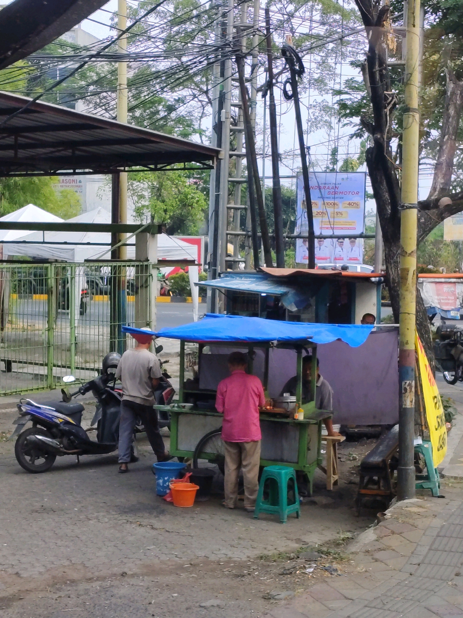 A street food vendor with a green cart and a blue canopy is operating on a street in Tangerang, Indonesia. A man in a pink shirt and khaki pants is standing behind the cart, seemingly preparing food. Another man in a gray shirt and dark pants stands to the left of the cart, with a blue bucket and an orange bucket nearby. A motorcycle is parked to the left of the vendor. In the background, there are tents, a fence, power lines, and trees. A billboard with text and images is visible behind the vendor's stall. The ground is paved with asphalt and some debris is present.