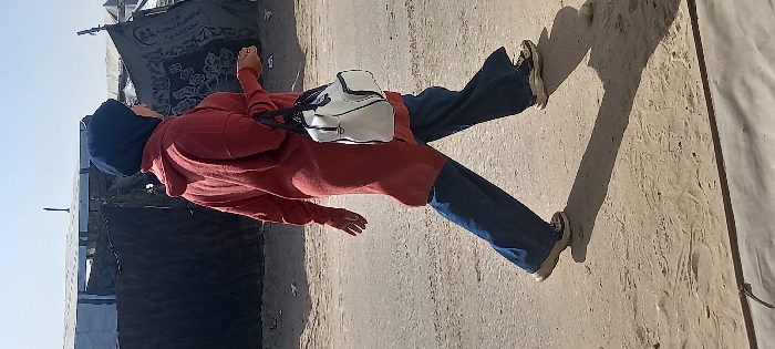 A person walks from left to right on a dirt and paved path in Khan Yunis, Palestinian Territory. The individual wears a dark blue hijab, a long-sleeved rust-colored tunic, blue wide-leg jeans, and light-colored athletic shoes. A white handbag with black accents is carried on their right arm. The person's head is slightly turned to the right. The background features several temporary shelters constructed from dark fabrics, tarpaulins, and possibly other materials, under a clear sky. One dark fabric structure displays white text and a patterned design.