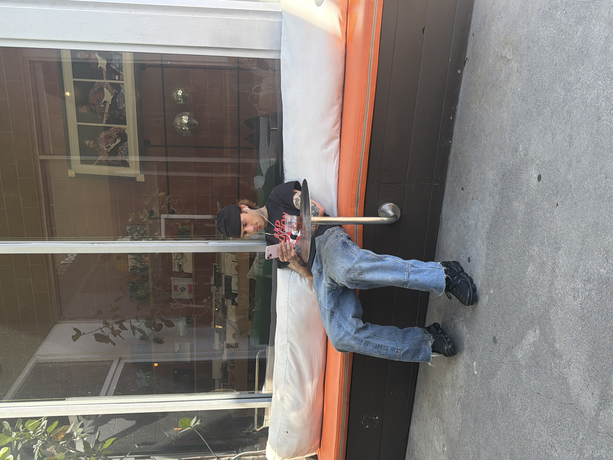 A full-length photograph captures a person seated on a built-in bench located outside what appears to be a cafe or restaurant. The individual, who is Caucasian, is wearing a black beanie, a dark t-shirt with a graphic print, light blue denim jeans, and dark-colored athletic shoes. The person is engrossed in using a pink smartphone. Visible tattoos cover the arms and portions of the neck.
The bench, where the individual is seated, has an orange-colored edge and a light-colored cushion. The bench is set against a dark-painted wall of the establishment. Behind the person and bench is a large storefront window reflecting the interior of the building and parts of the surrounding environment.
The scene seems to be set outdoors, on a concrete sidewalk or patio, adjacent to a building. The overall atmosphere suggests a daytime setting in an urban environment.
Although not explicitly stated in the image, based on the characteristics of the building, general architectural style, and foliage, one can speculate that the photograph was taken in the city of Los Angeles, United States of America.