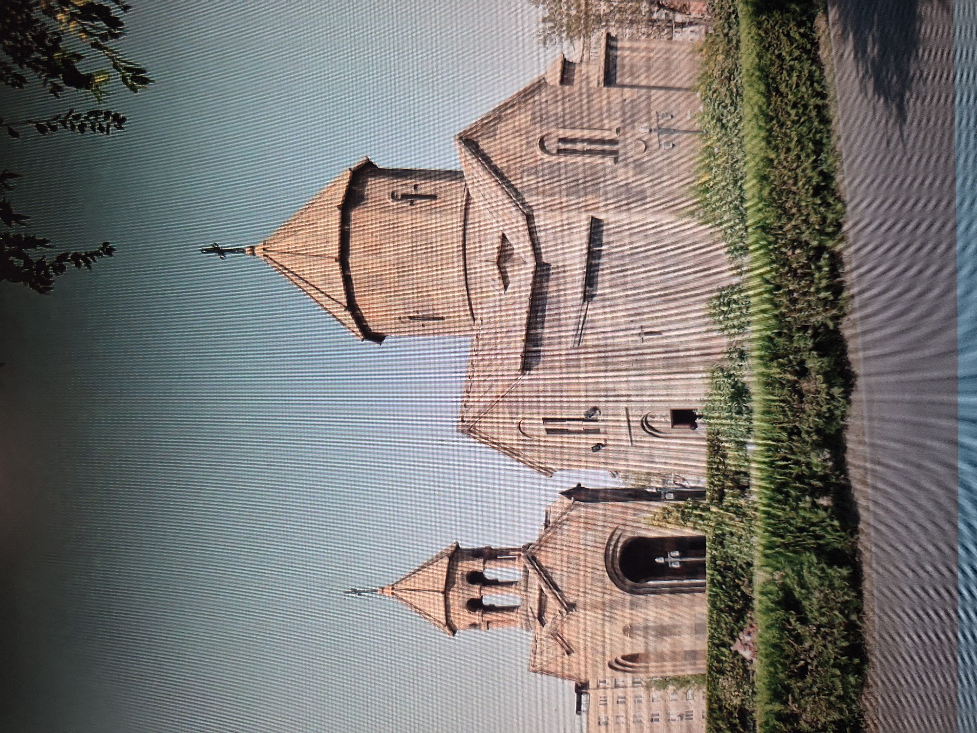 The image depicts a church located in Yerevan, Armenia. The building is made of reddish-brown stone and has a tiered structure with two prominent spires topped with crosses. The architectural style is indicative of traditional Armenian church design. The church is situated next to a neatly trimmed hedge and a paved area, possibly a walkway or road. The sky is clear and blue. Low-rise buildings are visible in the distance.