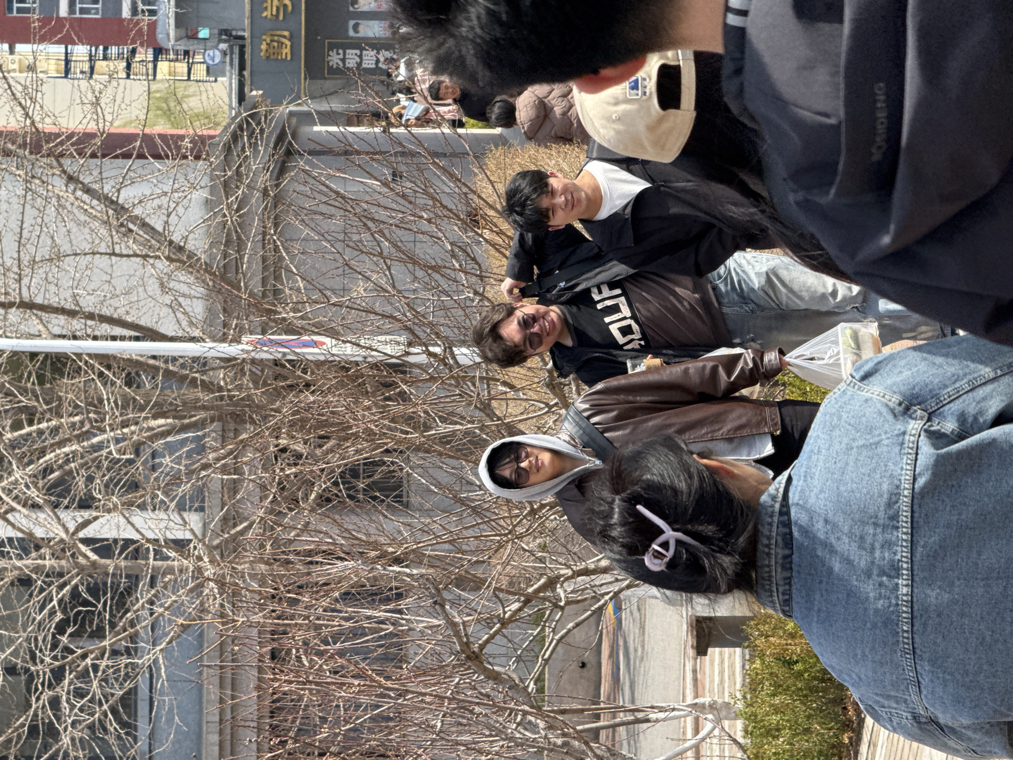 The image shows a group of people outdoors in Zhongtun, China. There are bare trees in the foreground. Several individuals are wearing casual clothing such as jackets and jeans. A person with sunglasses and a hood is visible. Another person is wearing a baseball cap. Some buildings are visible in the background. The overall environment suggests an urban or suburban setting during the cooler months.
