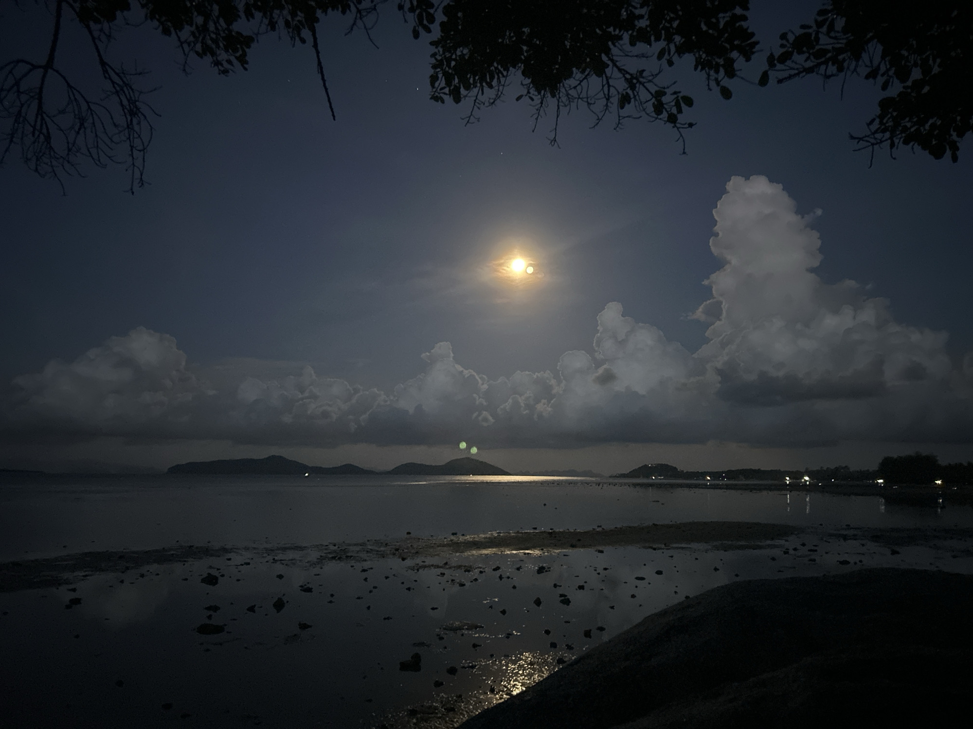 The image shows a night scene at a tidal flat in Ban Bang Khao, Thailand. The sky is a dark blue, with a bright moon obscured by light clouds. Tree branches frame the top of the image. The water is shallow and reflects the moonlight, creating shimmering highlights. In the distance, dark land masses are visible, possibly islands or hills. The foreground shows a dark, rocky tidal flat with scattered pools of water. No people are visible.