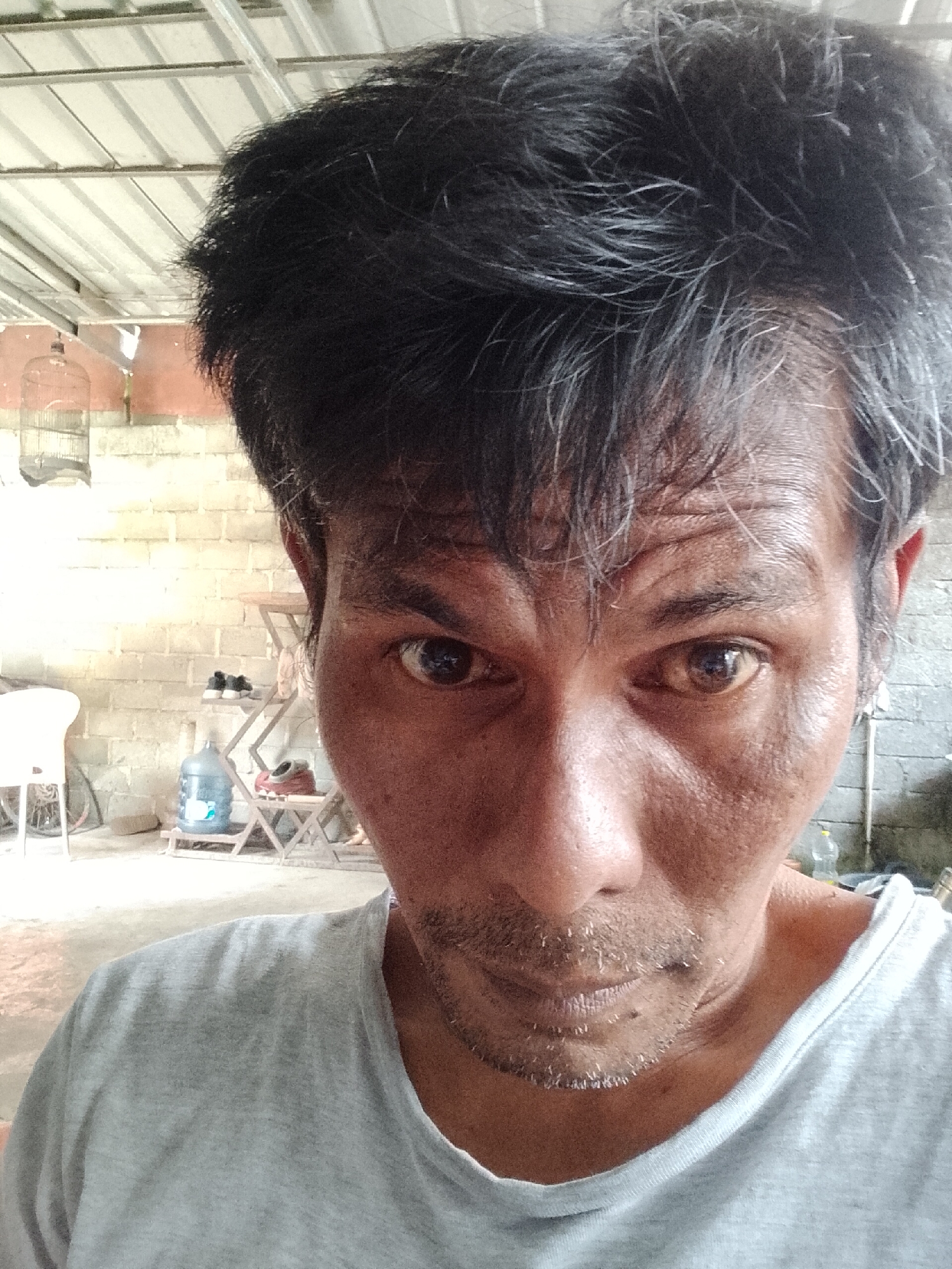 The image is a close-up selfie of a man in what appears to be an outdoor, possibly semi-rural, setting. He has dark, messy hair and is wearing a light gray t-shirt. His skin is tanned and appears to be sweating, with visible wrinkles on his forehead. He has a thin mustache and stubble. His eyes are dark and looking directly at the camera.
In the background, a wall constructed of light-colored bricks is visible. To the left, a birdcage hangs from a metal structure, and a white plastic chair is partially visible. Further back, there is a rack with items including a large blue water jug and a pair of shoes. The ground is a dirt or concrete surface. The overall impression is a candid, unposed shot taken in an informal environment. The context of Padang Dalom, Indonesia, suggests this could be a residential area or a common outdoor space within a community.