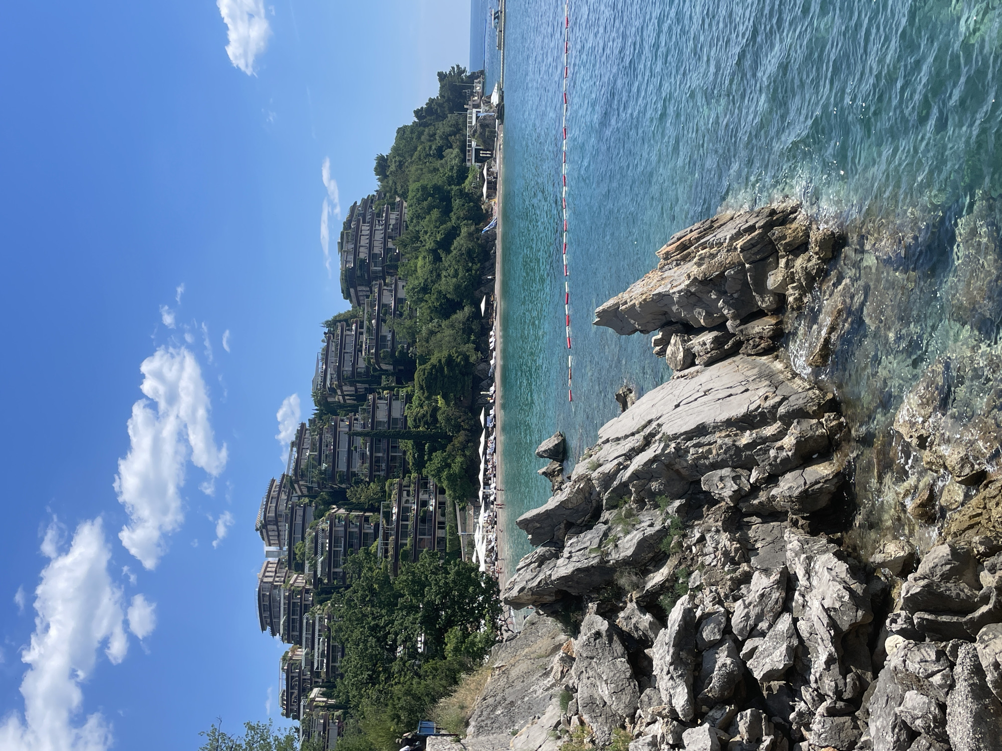The image shows a coastal scene in Budva, Montenegro. The view is from a rocky outcrop towards a beach and hillside.
The sea is clear and turquoise, with a red and white safety line visible. A sandy beach is populated with white sun umbrellas and people.
A steep hillside is covered with trees and multi-story buildings resembling apartments or hotels. The buildings are terraced and constructed in a manner that blends with the landscape.
The foreground is composed of large, grey rocks.
The sky is blue with scattered white clouds.