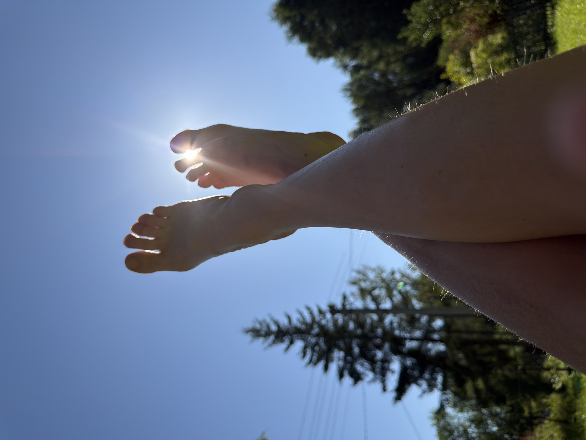 The image shows a pair of crossed legs and bare feet against a bright blue sky with the sun flaring between the toes. In the background, out of focus, are trees and power lines, suggesting an outdoor, possibly rural or suburban, setting. The overall impression is one of leisure and sunshine. The location is specified as Энтузиаст Снт (петрино), Russia.