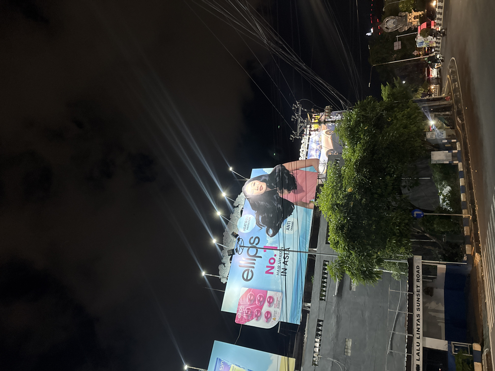 This media file captures a night scene in Denpasar Barat, Indonesia, featuring a large billboard advertising "ellips Hair Vitamin." The billboard displays a woman with long dark hair and the product packaging. The text on the billboard highlights "Heat & UV Protection" and "Anti Oxidant," along with the claim "No. 1 Hair Vitamin in Asia."
The setting appears to be a street, with overhead power lines, streetlights, and a road visible. The time of day is clearly night, as indicated by the dark sky and the illuminated billboard. The presence of a street sign reading "LALU LINTAS SUNSET ROAD" provides a specific location cue. A lush green tree is situated in front of a building behind the billboard, partially obscuring it. The overall mood is commercial and urban, with the bright billboard contrasting against the night sky.