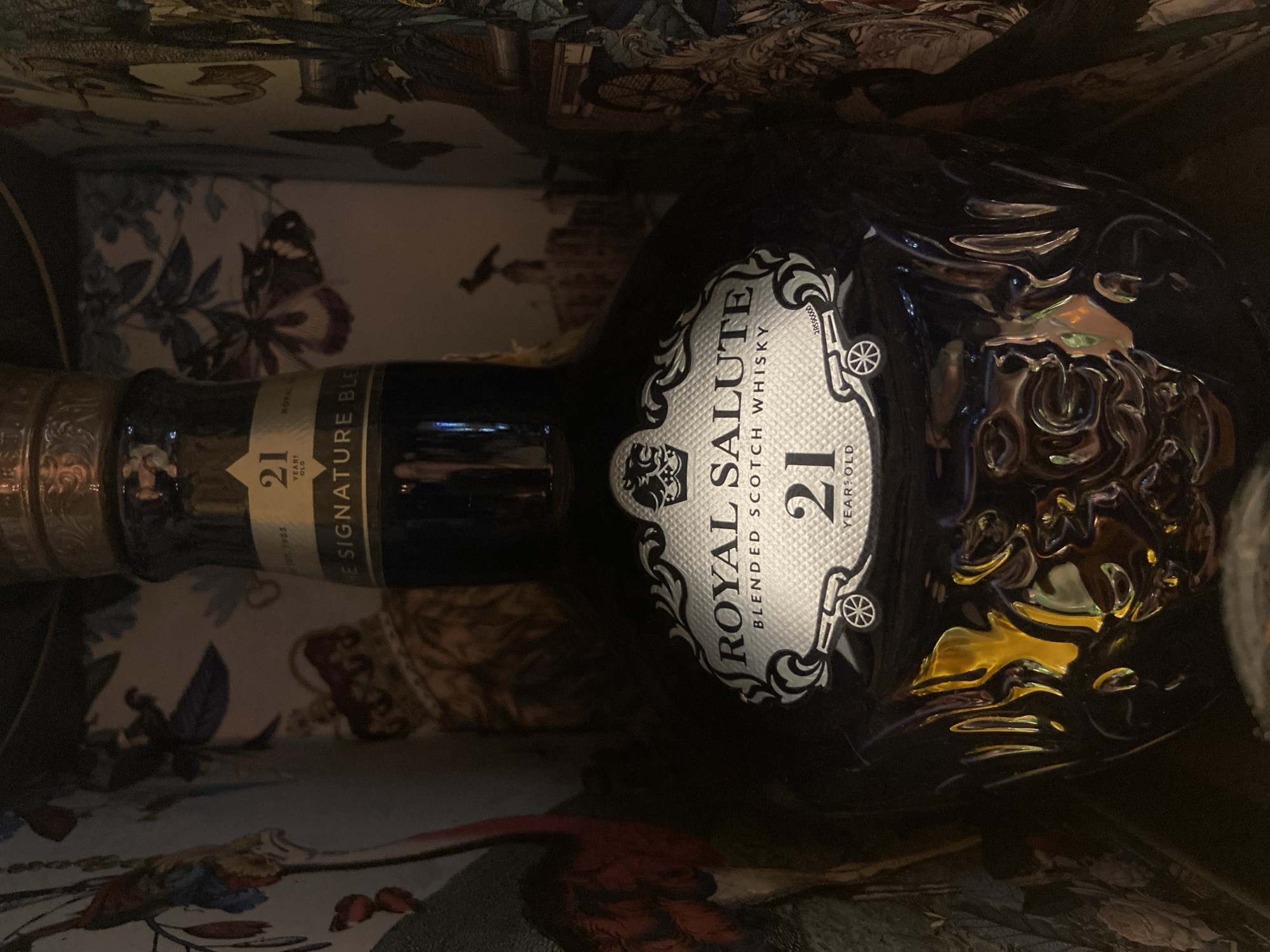 A close-up, slightly angled shot depicts a dark blue ceramic bottle of Royal Salute 21 Year Old Blended Scotch Whisky, nestled within a decorative box. The label prominently displays "ROYAL SALUTE BLENDED SCOTCH WHISKY 21 YEAR OLD" in silver on a white background with ornate filigree. The bottle is positioned horizontally with its neck to the left. The box interior features a patterned design with floral and butterfly motifs. The location is not apparent in the given image and cannot be determined.