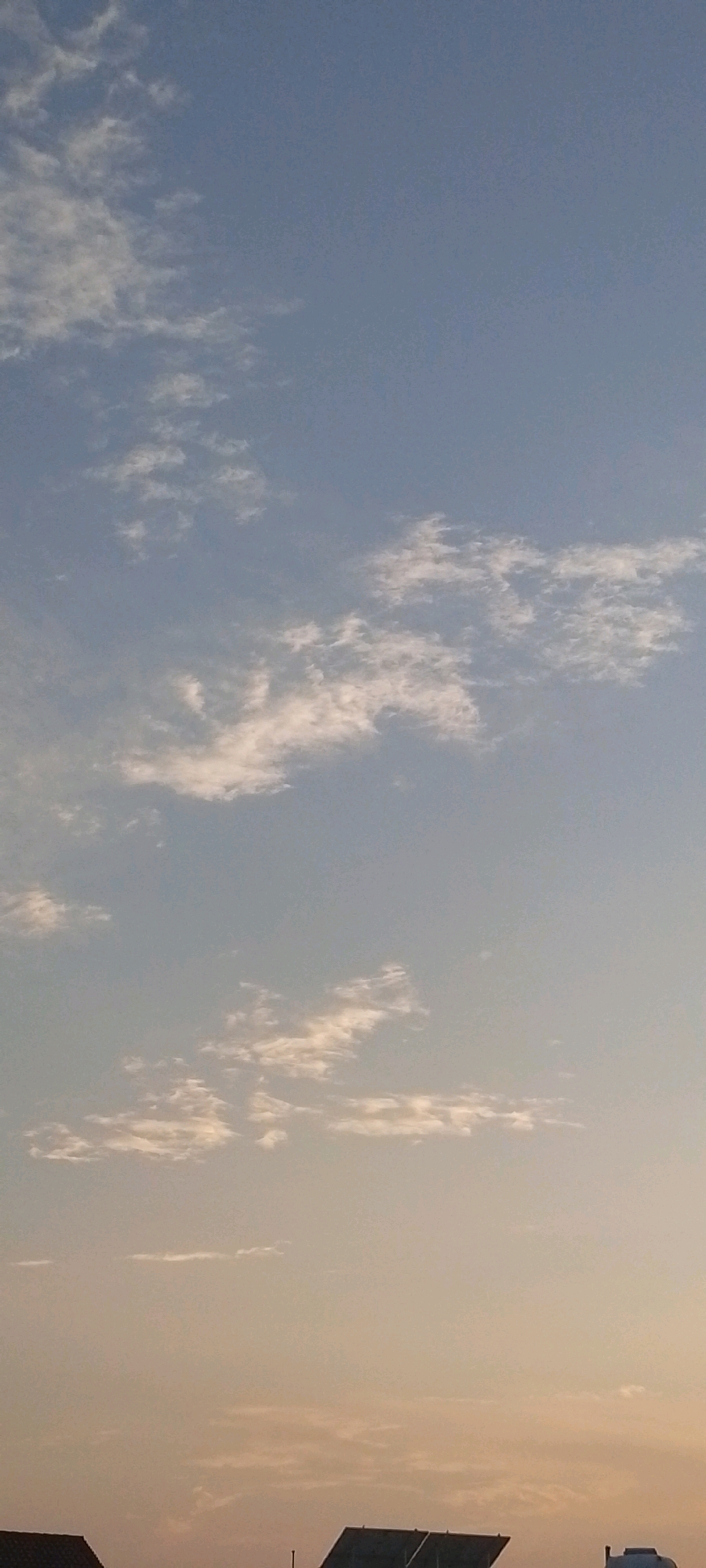 The image depicts a clear sky with scattered, wispy clouds during either sunrise or sunset, as indicated by the soft, warm hues present in the lower portion of the sky. The upper sky is a pale blue, gradually transitioning to a lighter, almost peachy color near the horizon. The clouds are sparse and appear to be high-altitude cirrus clouds.
In the foreground, the rooftops of a few buildings are visible, suggesting an urban or suburban environment. One building in the lower right corner has a large, dark, flat structure on its roof that could be solar panels or a similar installation. Another building to the left has a darker, pitched roofline. The overall impression is that of a peaceful, serene moment captured in Attock, Pakistan, likely at dawn or dusk. No people or specific actions are depicted.