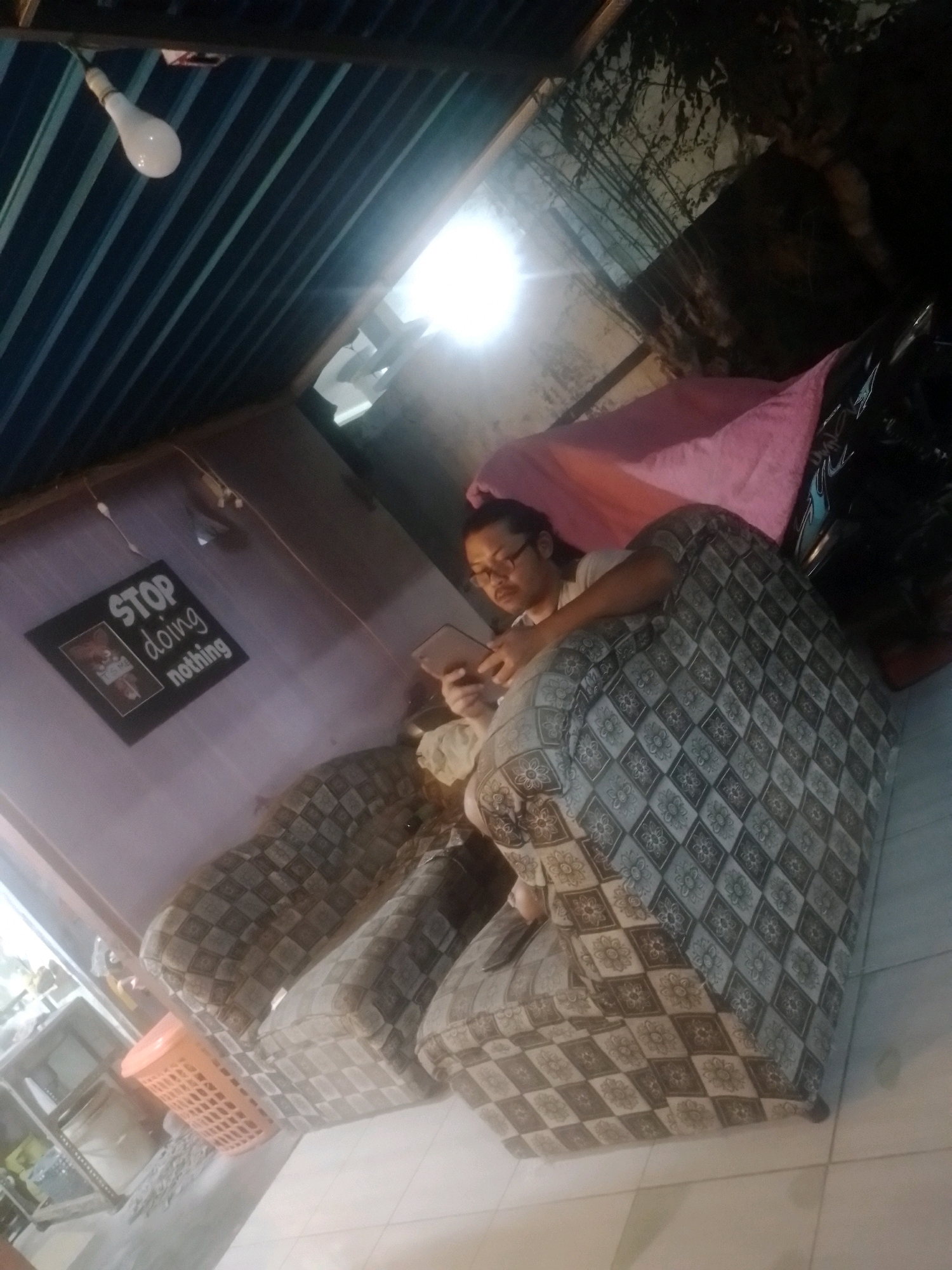 A person is sitting on a couch, holding a tablet. The couch has a patterned fabric with flowers and diamond shapes. A pink cloth is draped over another couch behind the person. On the wall, there is a framed picture with the words "STOP doing nothing" written on it. A light bulb hangs from the ceiling, illuminating the scene. The room appears to be an indoor space, possibly a living room, with a tiled floor. There is also a metal shelf with an orange basket on it in the background. The lighting suggests it is evening or night.