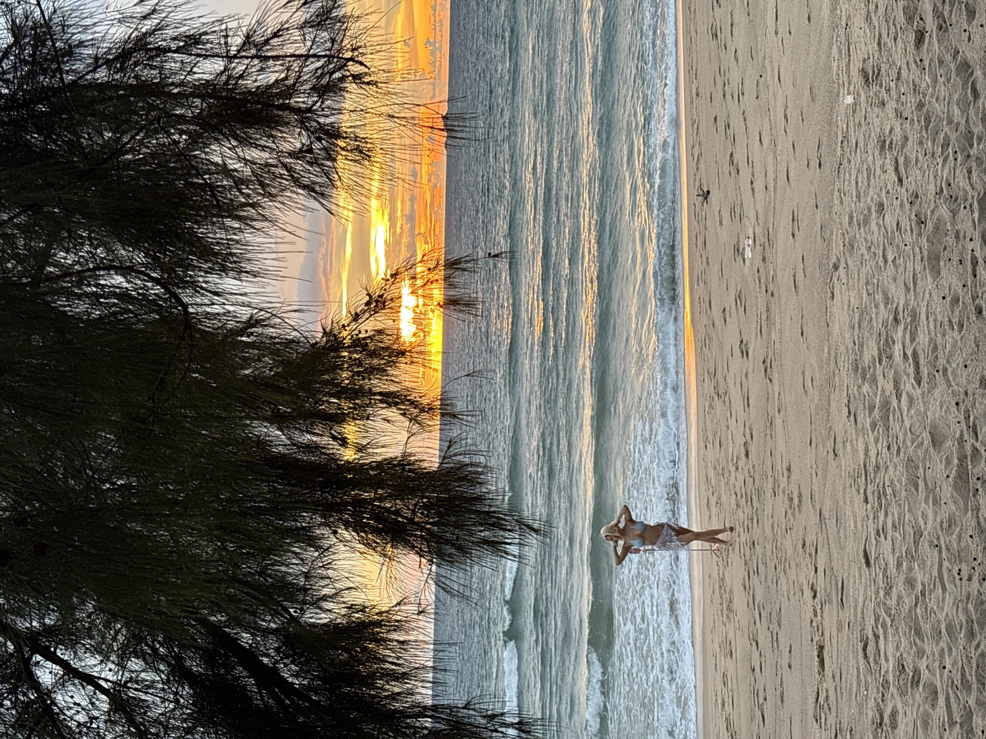 The image depicts a beach scene at sunset in Choeng Thale, Thailand, as evidenced by the tropical foliage framing the shot and the characteristic vibrant sunset over the Andaman Sea. In the foreground, a sandy beach is visible, textured with ripples and small dark specks, possibly dried seaweed or debris. A woman with blonde hair, wearing a light blue bikini and a sheer sarong, stands near the waterline, facing the ocean with her hands on her head. The ocean exhibits gentle waves breaking on the shore, reflecting the warm hues of the sunset. The sky is a dramatic display of orange, yellow, and pink clouds, with the sun partially obscured by them and a silhouette of a pine-like tree hangs over the scene. The overall atmosphere is serene and picturesque.