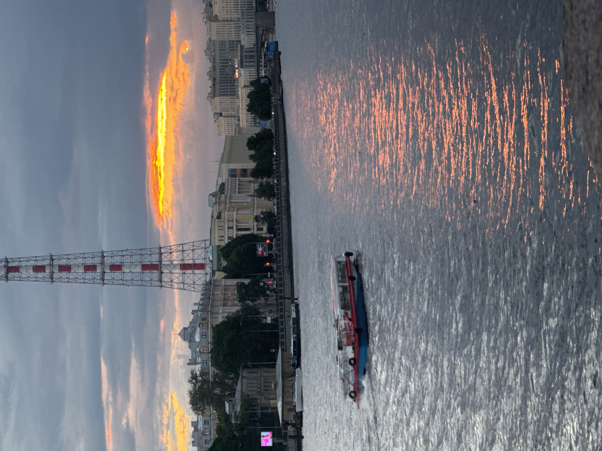 This is a medium-distance, eye-level, vertical shot of the Neva River in Saint Petersburg, Russia, during sunset. In the foreground, a red and blue passenger boat is moving from left to right, creating a wake. The river's surface reflects the warm orange and yellow hues of the sunset, contrasting with the darker, choppy water. In the mid-ground, along the riverbank, are several buildings with grey roofs and white facades, interspersed with trees. Further back, a tall, red and white lattice tower dominates the skyline. The sky is overcast with dramatic clouds, with a bright band of orange and yellow light peeking through them, indicative of the setting sun. The overall scene captures a moment of twilight in an urban riverine environment.