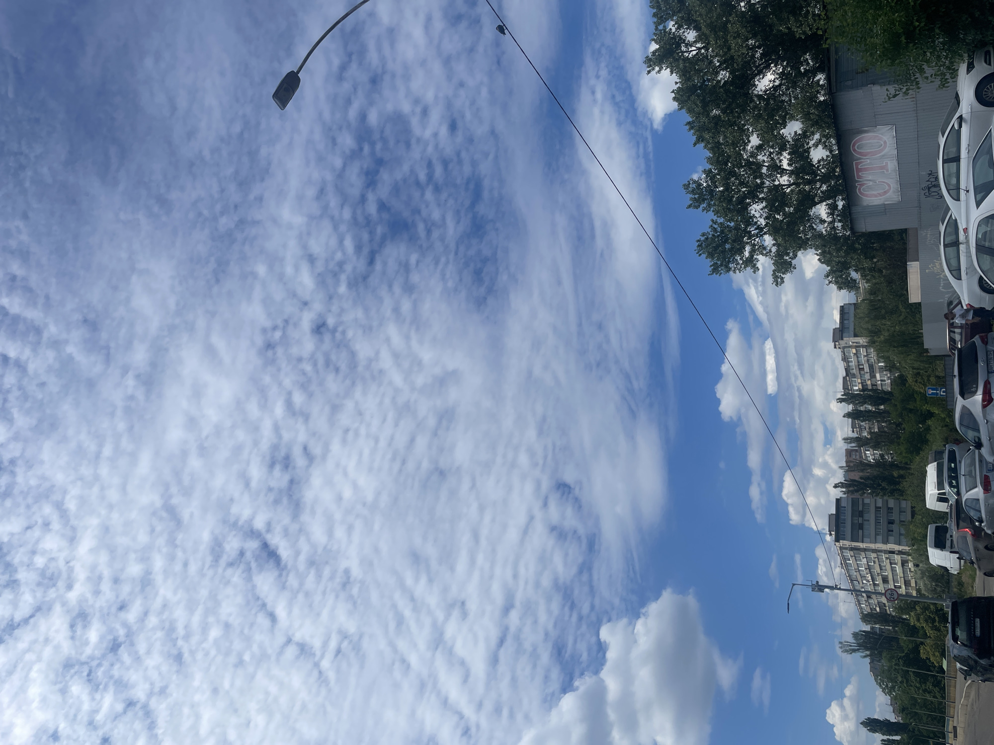 This is a photograph taken from a street level perspective, looking upwards towards a partially cloudy blue sky. In the foreground, several cars are parked or driving on a road. Beyond the cars, there is a row of apartment buildings and dense green trees. A streetlamp is visible on the right side of the frame, and a power line stretches across the image. On a corrugated metal structure in the mid-ground, a sign with the letters "СТО" in red is visible. The image appears to be taken on a bright day. While the specific location is not definitively identifiable, the architecture of the apartment buildings suggests an urban environment, and the presence of the Cyrillic letter "СТО," commonly used for "Service Technical Operations" in Ukraine and other post-Soviet countries, strongly suggests the location is in Kyiv, Ukraine. There are no people clearly visible interacting with each other.
