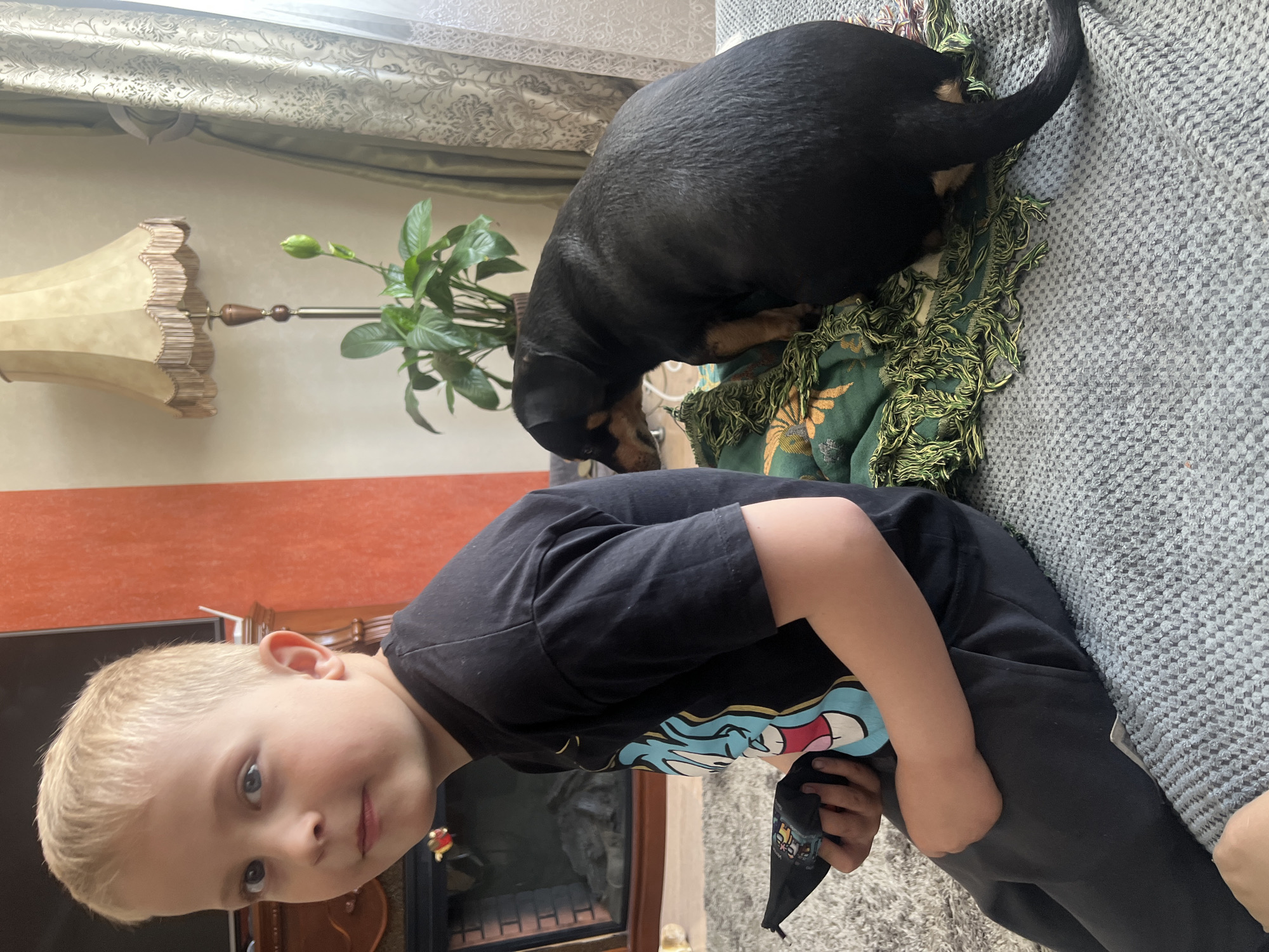 A photograph shows a young boy and a dog on a gray couch. The boy, with blonde hair and wearing a black t-shirt, is in the foreground. A black and tan dog is resting on a green fringed blanket on the couch behind the boy. In the background, there is a floor lamp, a potted plant, and a wall painted in orange and tan.