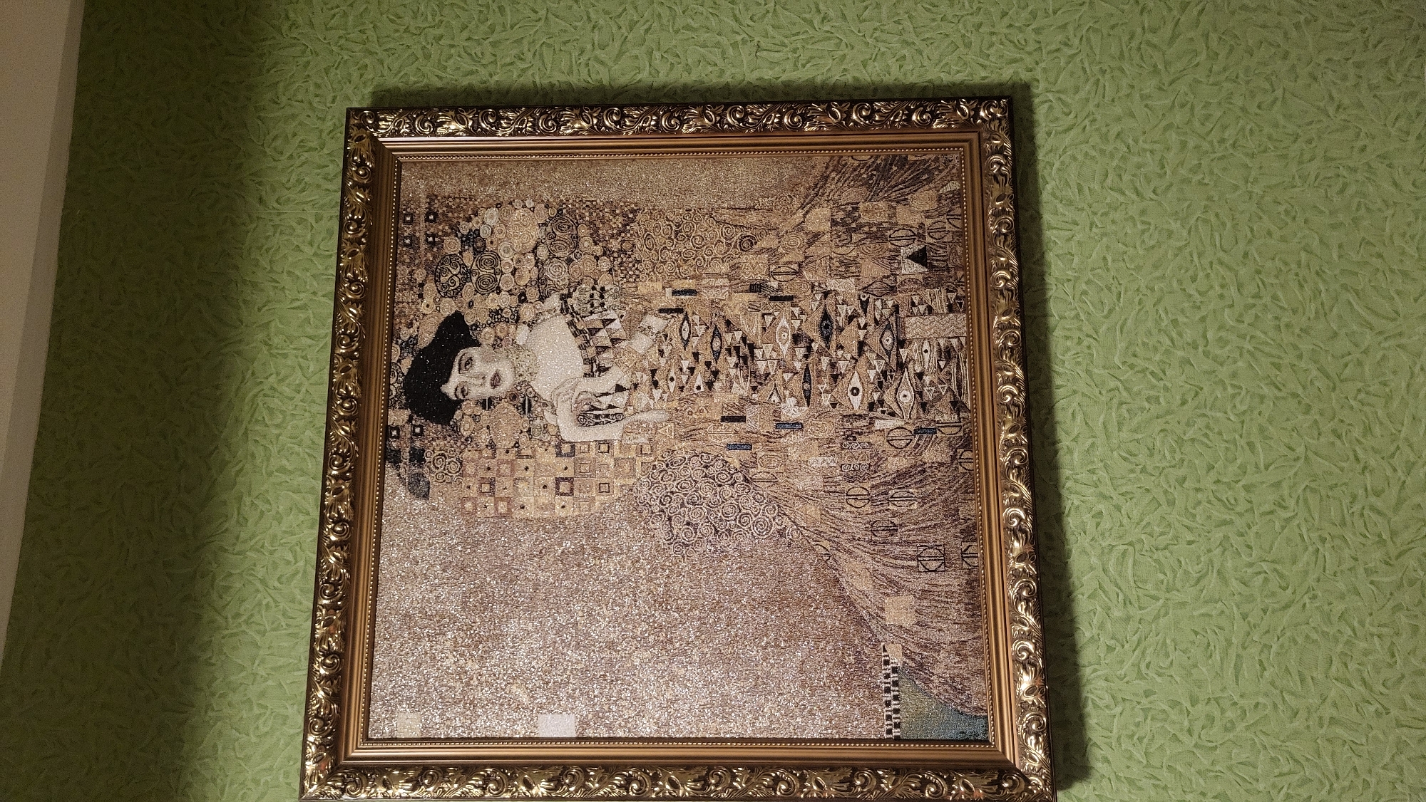 A framed artwork, likely a mosaic or tapestry depicting Gustav Klimt's "The Kiss," is hung on a textured, light green wall. The artwork features a couple embracing, rendered in a style characterized by elaborate patterns and gold tones. The frame is ornate and gold-colored. The background wall has a subtle, repeating organic pattern. The scene is indoors, suggesting a domestic or public space, and the lighting appears to be ambient. There are no people visible in the frame other than those depicted within the artwork. The overall impression is one of art appreciation and home decor. The image was taken in the city of Smila, Ukraine.