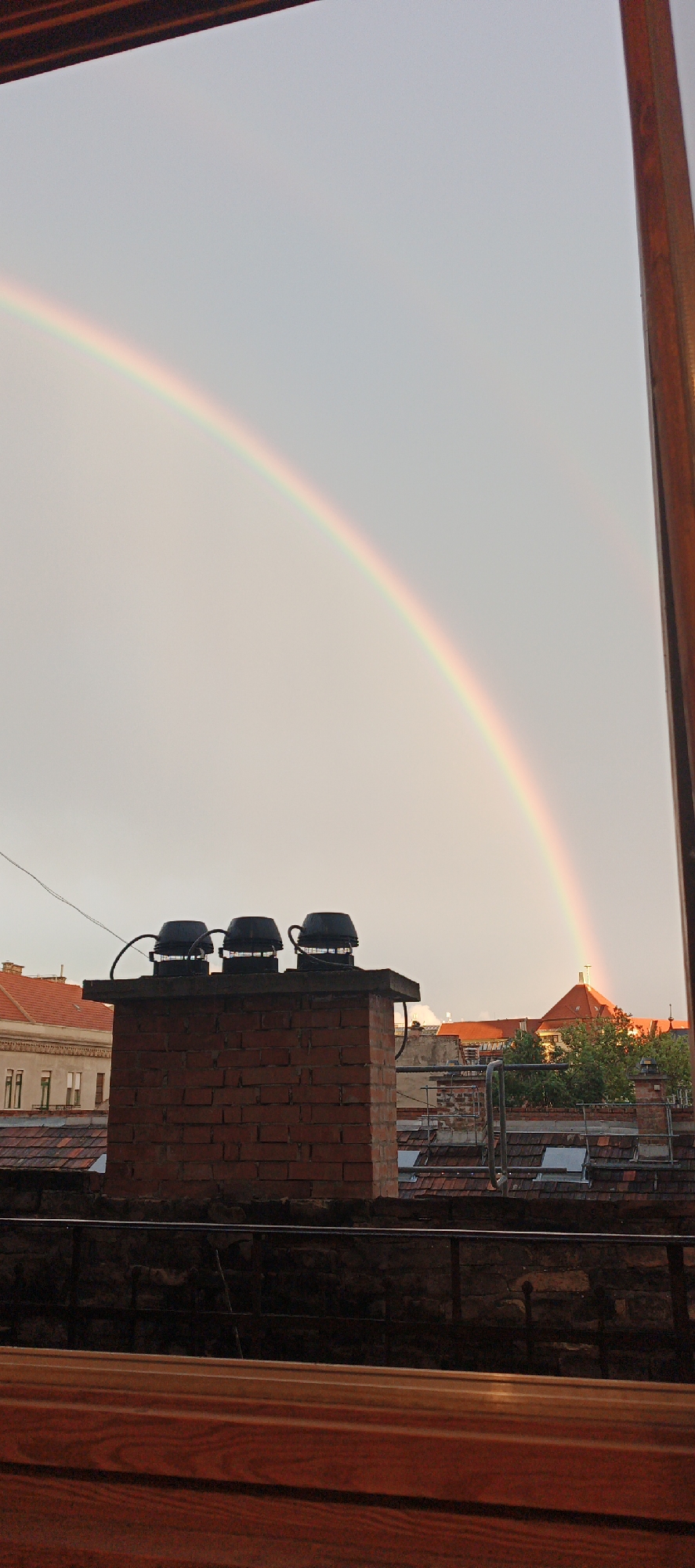 A view from a balcony or window overlooks the rooftops of Budapest, Hungary, showcasing a vibrant rainbow against a pale sky. In the foreground, a brick chimney with three black vent caps stands prominently. Additional rooftops with reddish-brown tiles fill the middle ground, along with glimpses of trees. Elements of the balcony structure partially frame the scene.