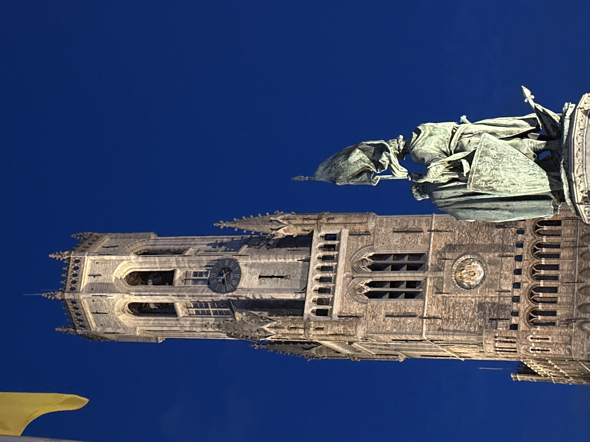The image shows the Belfry of Bruges, a medieval bell tower located in the city of Bruges, Belgium. The tower is made of brick and stone and is topped with a spire. In front of the tower is a statue of two men, one of whom is holding a flag. The sky in the background is a deep blue, suggesting that it is nighttime. The tower is illuminated, casting a warm glow on the surrounding area. A yellow flag is visible in the upper left corner of the image.