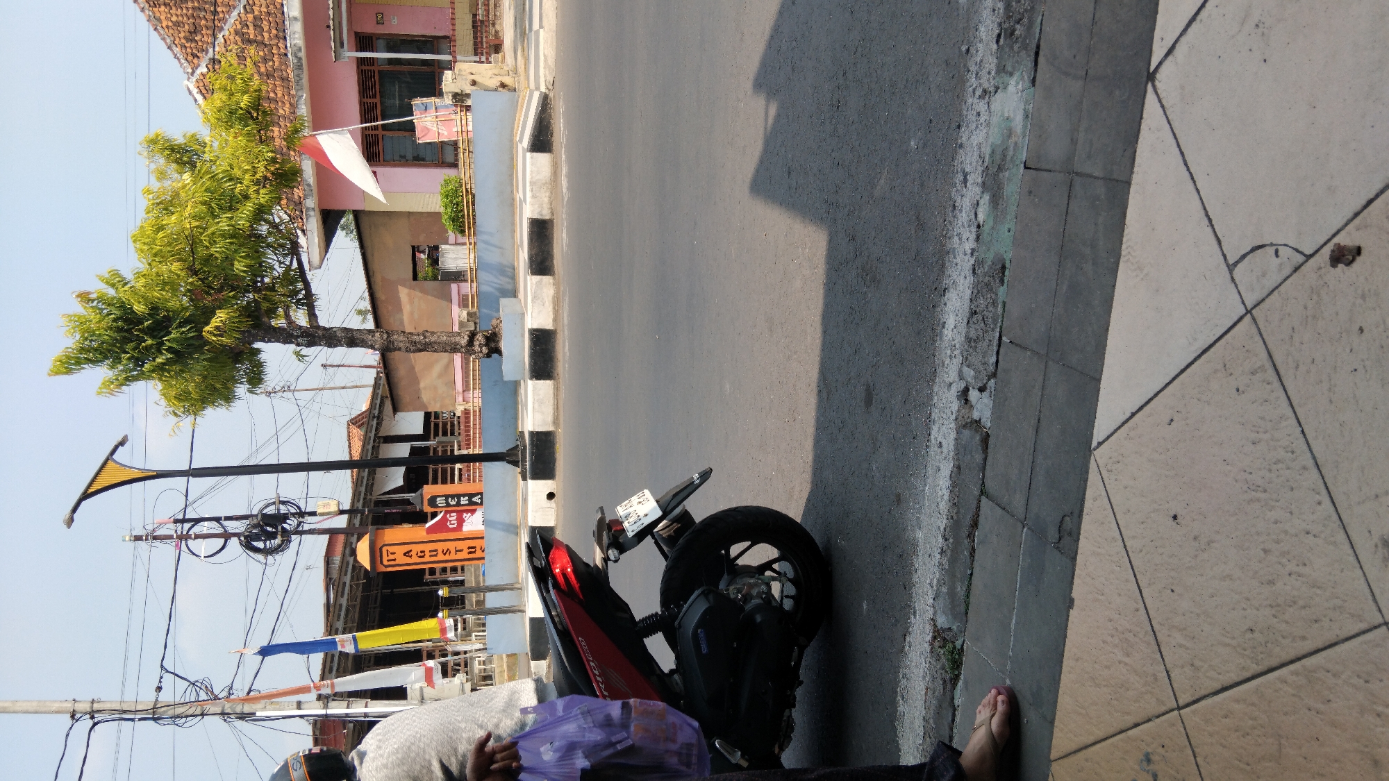 An outdoor, eye-level, full shot captures a scene on a paved road in Batang, Indonesia. In the foreground, a red and black motorcycle is parked on the asphalt. A person, partially visible on the left, is holding a purple plastic bag. The background features buildings with tiled roofs, a tall tree with green foliage, and utility poles with electrical wires. A flag, red and white, is displayed on one of the buildings. The road has a curb with black and white markings. The overall environment appears to be a street with commercial or residential buildings lining the road.