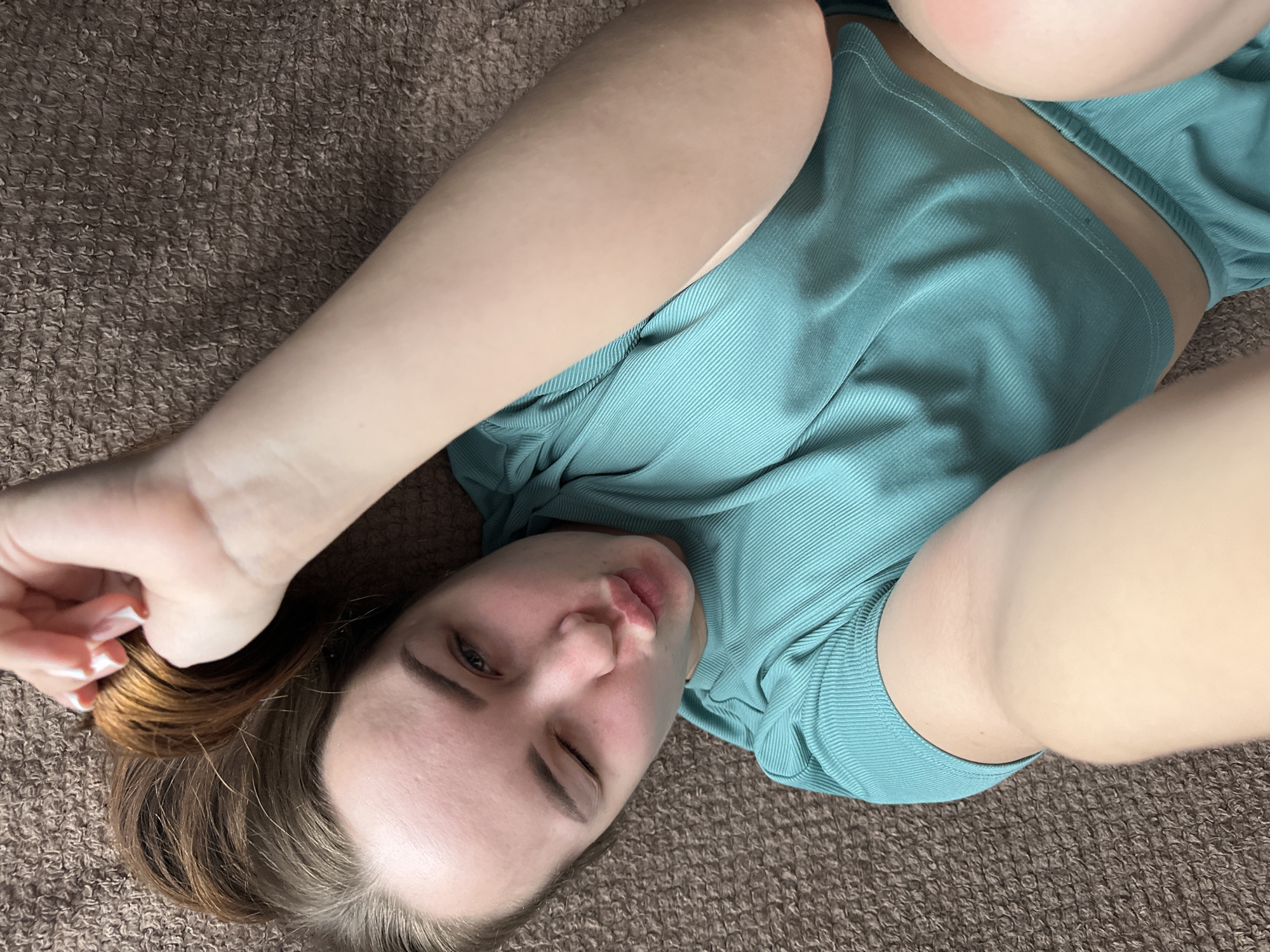 A woman with light skin and brown hair lies on a brown, textured surface, possibly a bed or carpet. She is wearing a ribbed, teal-colored short set. She is winking with her right eye and has her arms raised, with one hand near her head. The background is the same brown texture. No indication of Chaikovsky, Russia, or city elements are present in the image.