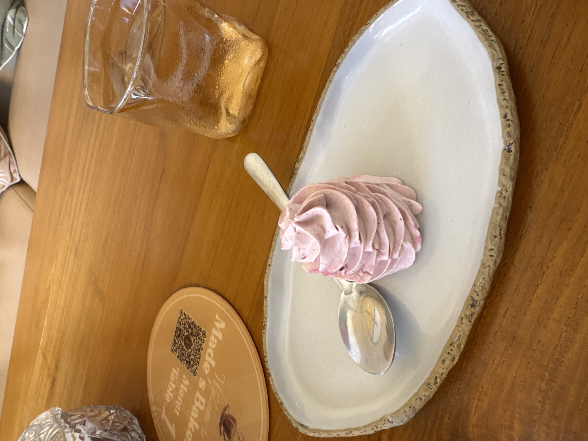 A top-down shot depicts a table setting in Kuta Selatan, Indonesia. The table is wood. A clear glass pitcher filled with amber liquid is in the top left corner. Next to it is a plate with a light pink, swirled dessert, served with a spoon. Adjacent to the plate is a circular coaster labeled "Made's Baker Menu Table". In the bottom left is a decorative container.