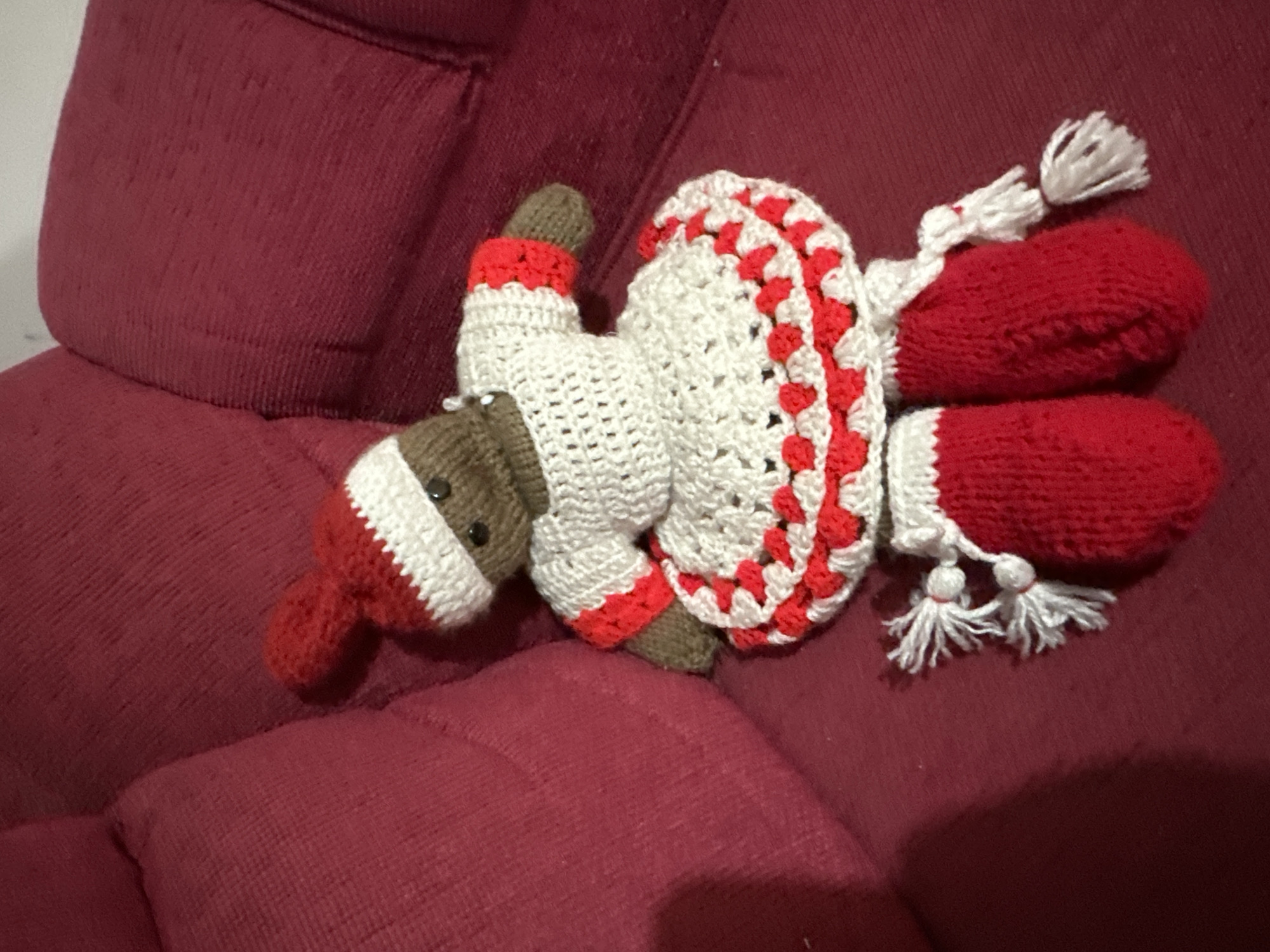 A crocheted toy doll is lying on a red upholstered couch. The doll has a brown head and feet, a white body, red appendages, and white tassels. The scene appears to be indoors, but the location in the city of Arica, Chile, cannot be determined from the image.
