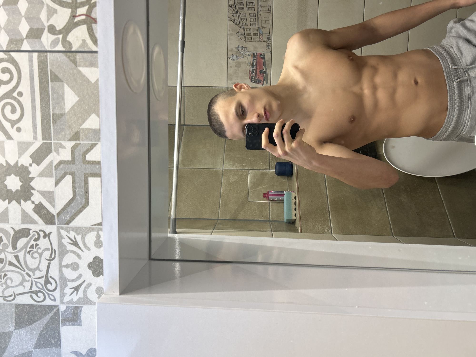 The image is a mirror selfie taken by a young man in what appears to be a bathroom or shower area. The photo is oriented vertically but the subject appears rotated counter-clockwise.
The primary subject is a shirtless young man with a buzz cut hairstyle. He is physically fit, displaying visible abdominal muscles, defined chest, and shoulders. He holds a black smartphone in his right hand, using it to take the mirror selfie. His facial expression is neutral. He is wearing grey sweatpants or shorts, visible from the waist down.
The setting is an indoor space, likely a bathroom, with various types of tiles. The wall directly behind the man's reflection features light brown or beige square tiles. Above these, there is a decorative border with a cartoon-like illustration of European-style buildings and a red double-decker bus. The floor in the reflection is also tiled with similar light brown/beige squares. To the left of the mirror, outside the reflection, is a wall covered in grey and white patterned tiles, featuring intricate geometric and floral designs.
Inside the reflected shower area, a vertical silver pole (possibly part of a shower rail or structure) is visible on the left. On the floor behind the man's reflection, a pink bottle and a blue/green sponge can be seen. A white, circular, shallow basin or tray is visible near the man's lower body, possibly a shower tray or toilet base. The mirror itself has a white or light grey frame, and two circular, slightly raised white objects are visible on the wall to the top right of the mirror's edge.
The activity is a self-portrait (selfie) taken by the man in a mirror. There is no visible text. The location context provided is Burgas, Bulgaria, which aligns with the European-style tile patterns and illustrations. The time of day is not discernible from the indoor lighting.