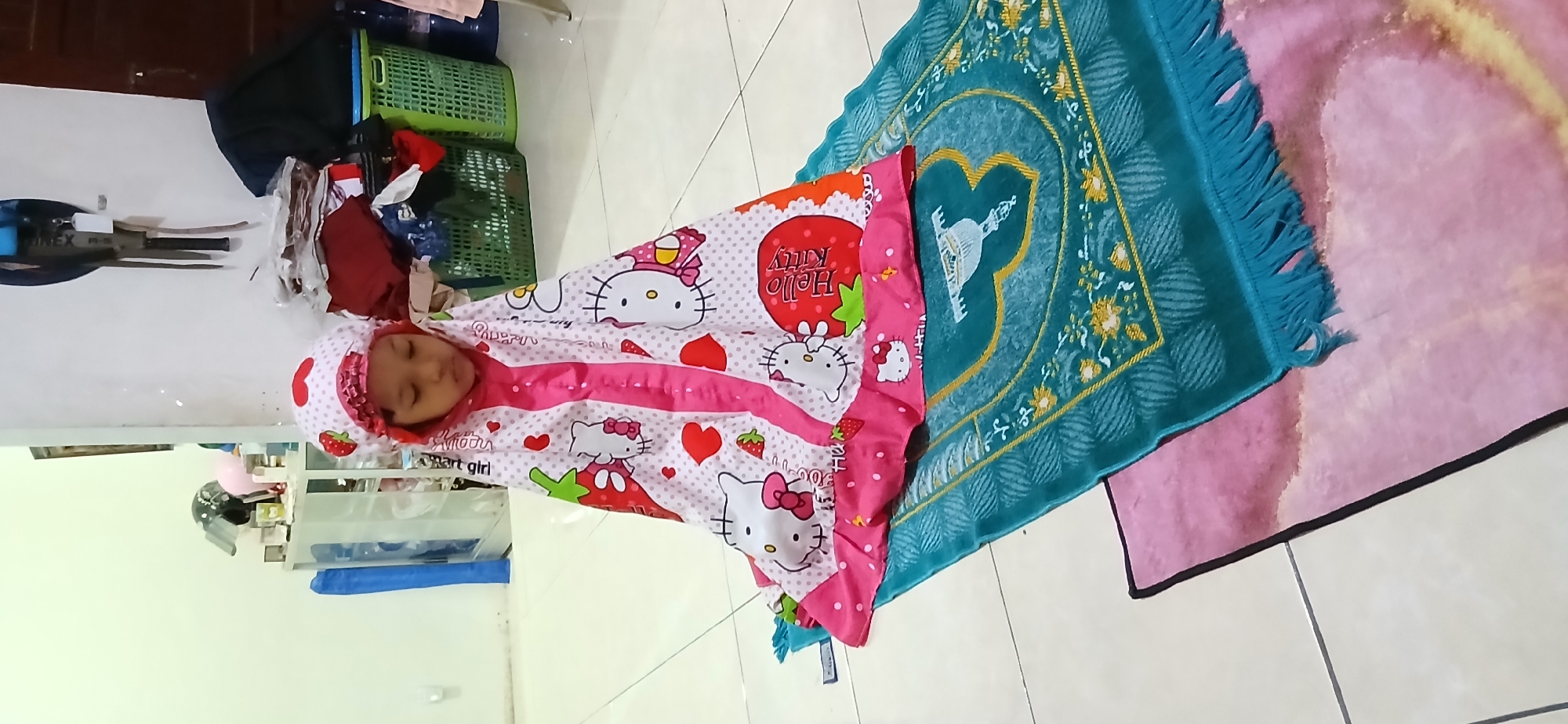 A young girl is kneeling on a prayer mat in what appears to be an indoor setting. The girl is wearing a pink hijab with white polka dots and a strawberry pattern, and a white prayer dress adorned with Hello Kitty characters and strawberries. The prayer mat is teal with intricate gold and white patterns, including a depiction of a mosque. Beneath the teal mat is another pink mat. The background shows a tiled floor, a white cabinet with various items, and laundry baskets filled with clothes. The overall environment suggests a domestic space, possibly a home or a room within a building in Samarinda, Indonesia.