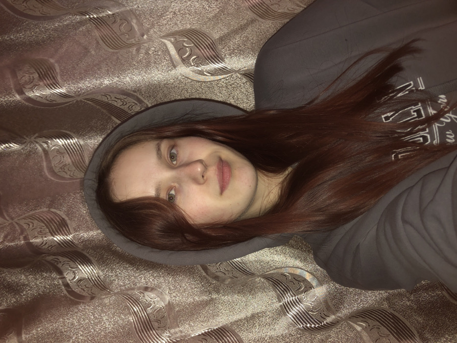 A single female individual is centrally depicted in a medium close-up, looking directly forward. She has long, reddish-brown hair, light-colored eyes, and fair skin, exhibiting a neutral expression with a slight smile. She is wearing a grey hooded sweatshirt with the hood drawn up, covering her head. Partially visible white lettering on the sweatshirt reads "BROOKLYN new York." The background is an indoor setting featuring a curtain or fabric with a patterned design of wavy, vertical elements and intertwined ornate shapes in a light brown or beige color. The lighting appears to be artificial. Based on the provided information, the location is the city of Фоки, Russia.