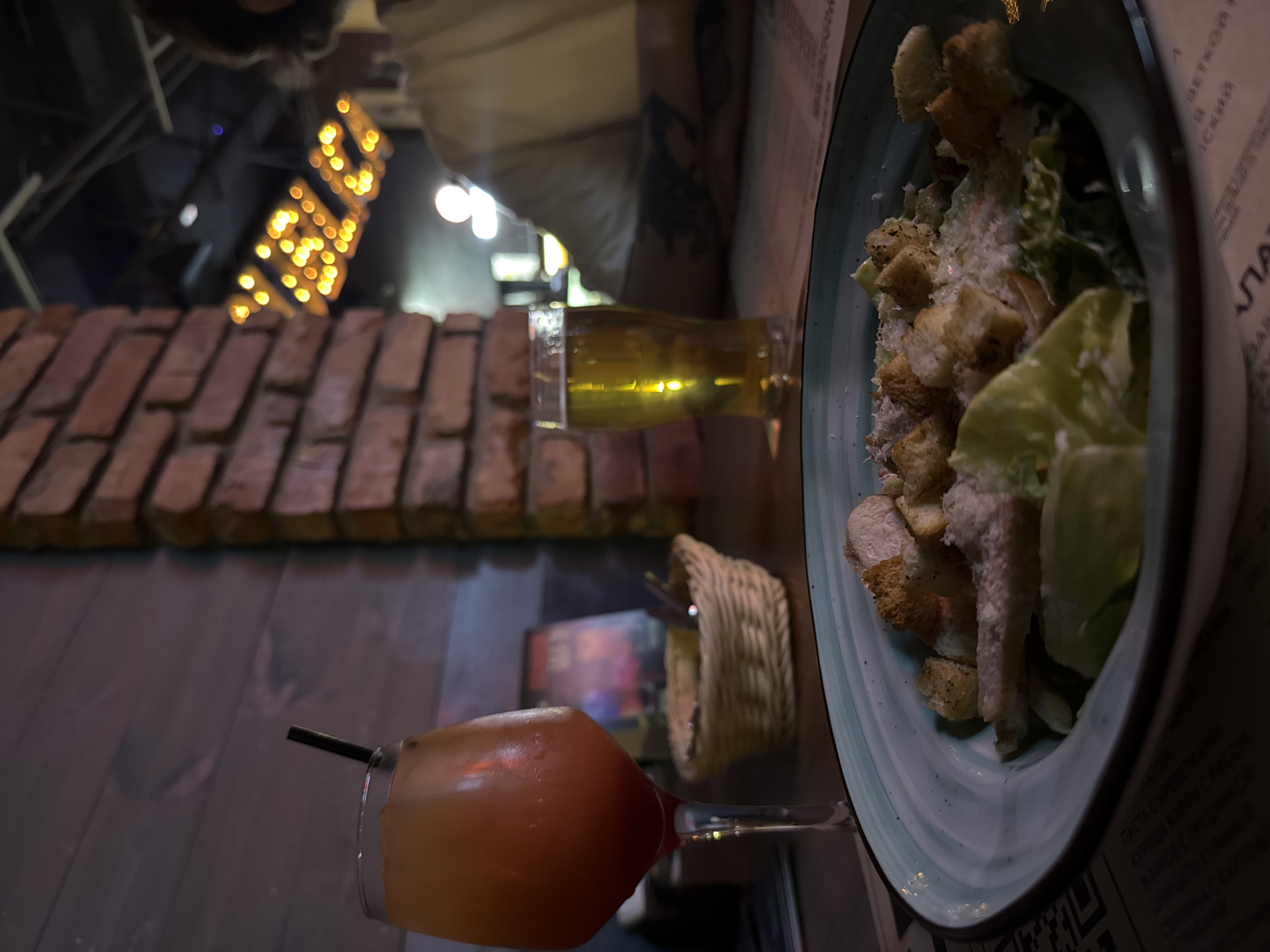The image depicts a close-up view of a table at what appears to be a restaurant or bar. The primary subjects are food and drinks. In the foreground, a glass of amber-colored liquid, likely a cocktail or beer, sits on the table, complete with a dark straw. Adjacent to this is a plate of Caesar salad, featuring lettuce, croutons, and possibly chicken or fish, sprinkled with grated cheese. A small woven basket is also visible.
In the background, a brick wall element with decorative lighting resembling strings of yellow bulbs suggests an interior setting. The lighting is dim, creating a cozy and intimate atmosphere, and the overall impression is that of a late evening or nighttime scene. The image is rotated 90 degrees clockwise.
There is visible text on a paper menu or placemat near the salad, but it is mostly illegible due to the focus and angle. Some Cyrillic characters are discernible, hinting at a Russian-speaking location, consistent with the provided context of Novosibirsk, Russia. No people are clearly visible in the frame, though the presence of food and drinks implies a dining or social occasion.